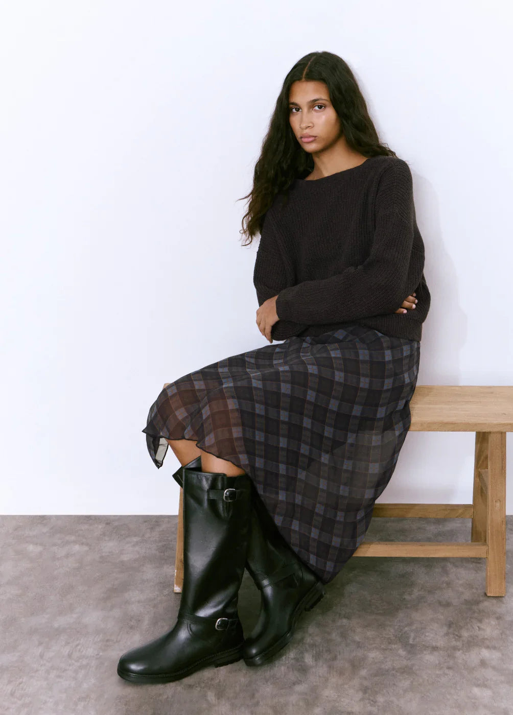 אישה יושבת על ספסל בחולצה סרוגה כהה, חצאית משובצת ומגפיים שחורים, אופנה CHOZEN