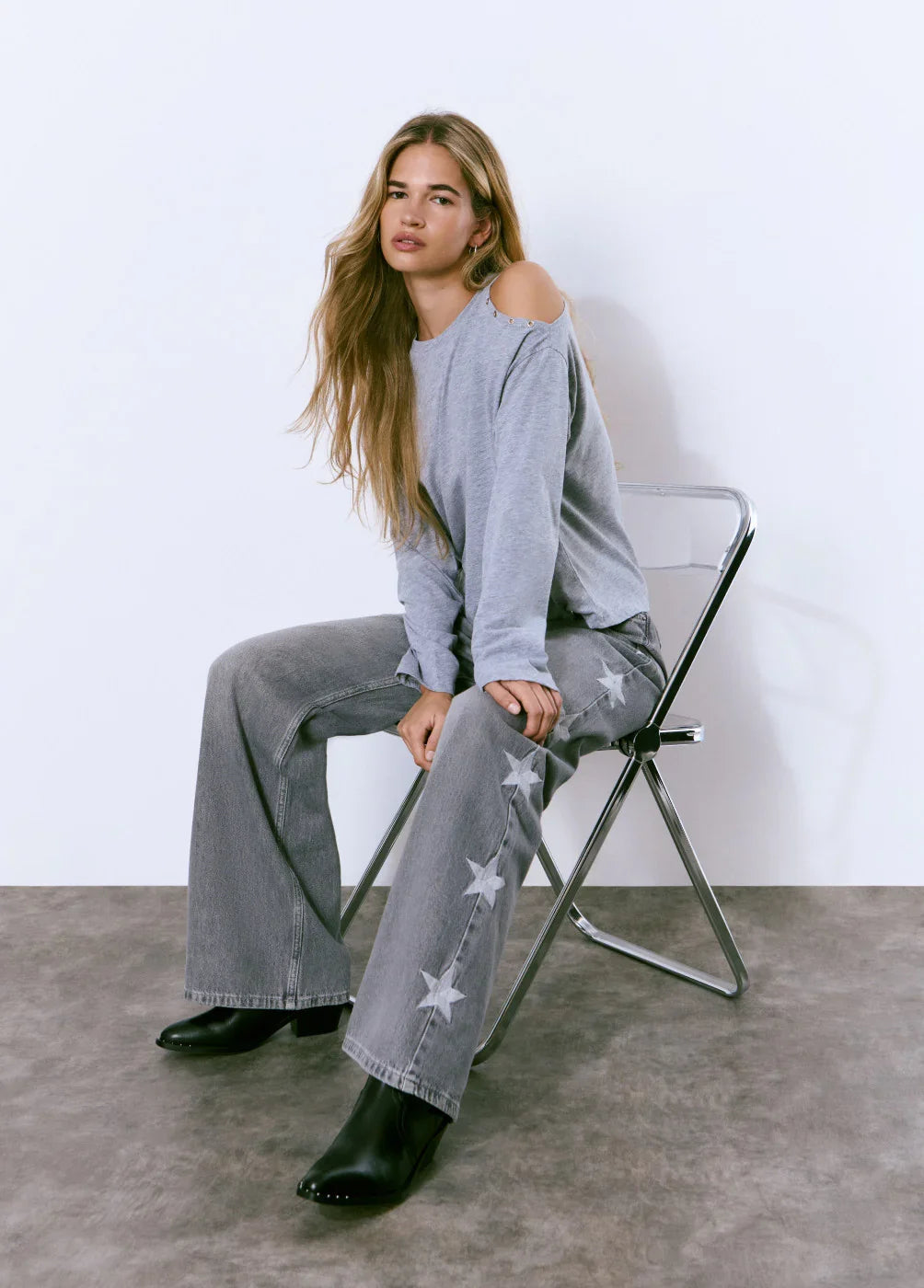 דוגמנית יושבת על כיסא, לובשת חולצה אפורה עם כתף חשופה ומכנסי ג'ינס אפורים עם כוכבים, אופנה טרנדית CHOZEN