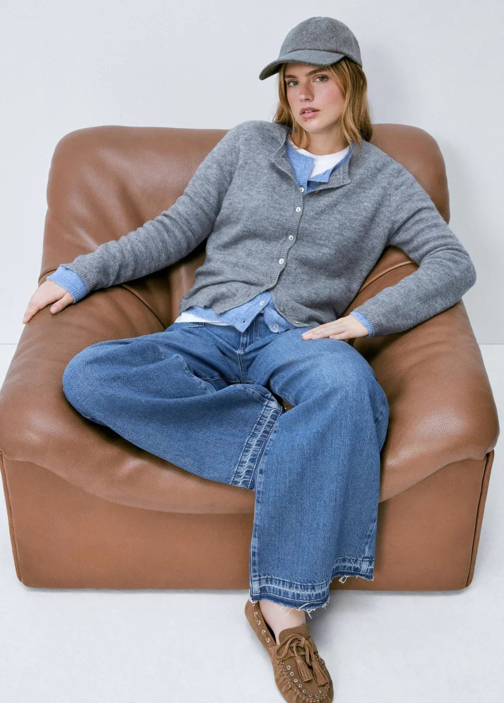 אופנת נשים CHOZEN, דוגמנית עם סוודר אפור, ג'ינס רחב וכובע, יושבת על כורסה חומה