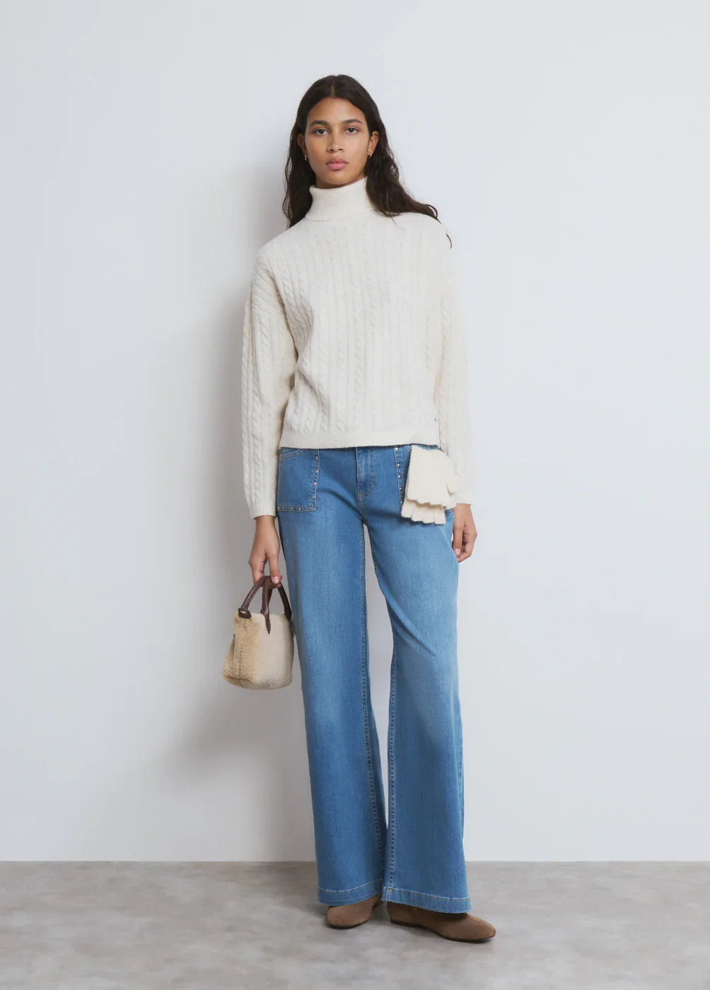 אישה לובשת סוודר לבן עם גולף, מכנסי ג'ינס רחבים, נעלי זמש ותיק יד אופנתי. בגדי חורף CHOZEN