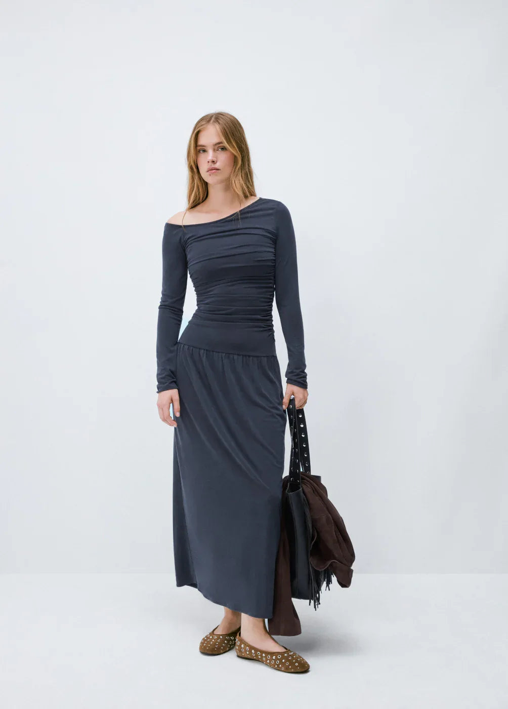 אישה לובשת שמלת מקסי כחולה כהה עם שרוולים ארוכים ועקב אחד חשוף, נושאת תיק אופנתי – CHOZEN