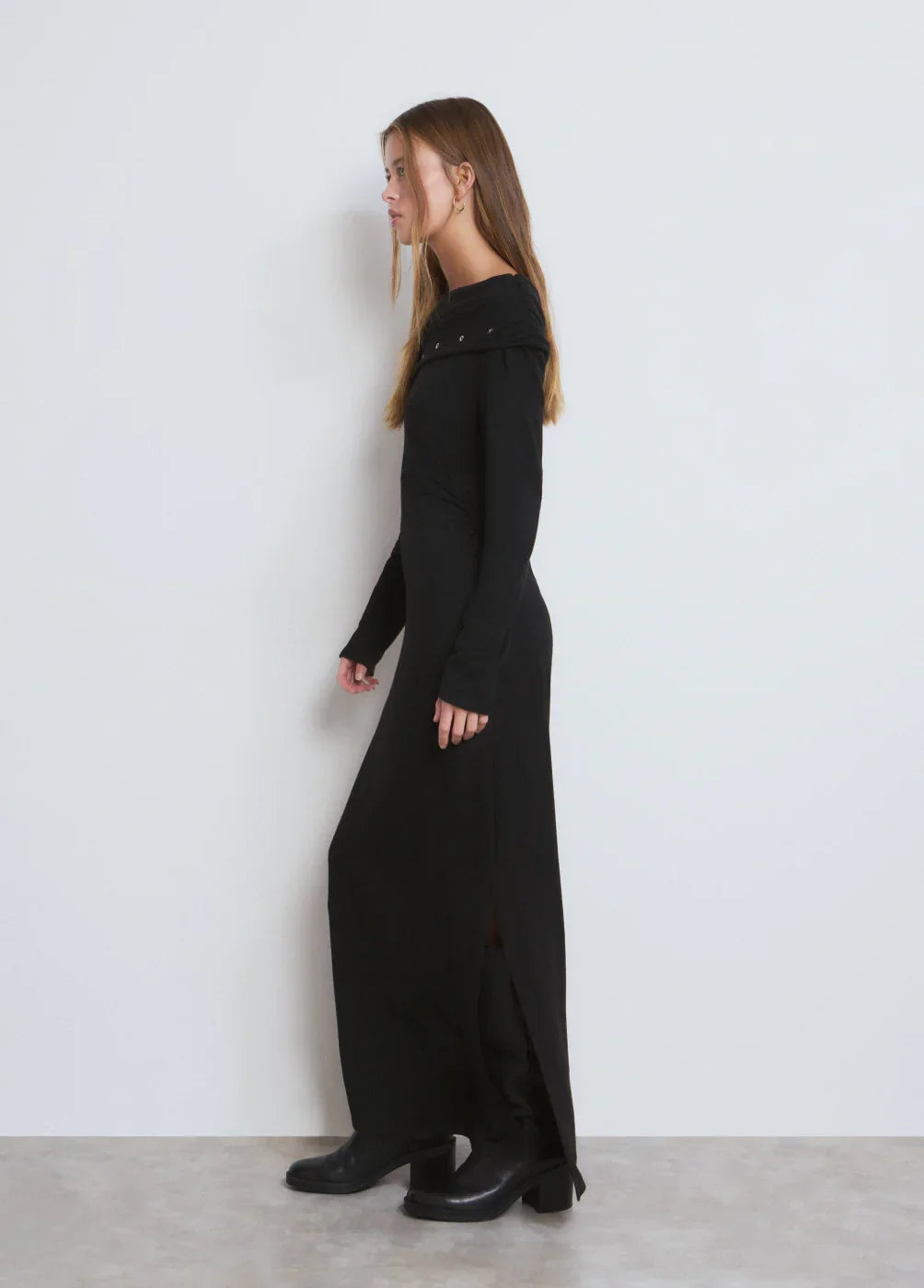 דוגמנית עם שמלה שחורה ארוכה ומגפיים, על רקע קיר לבן, אופנה לנשים CHOZEN