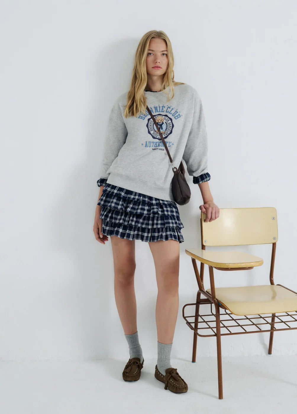 דוגמנית לובשת סווטשירט אפור עם כיתוב וחצאית משובצת כחולה, תיק קטן, יושבת על כיסא בבית CHOZEN