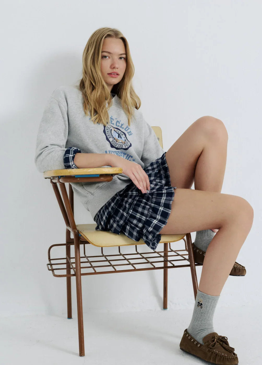 דוגמנית יושבת על כיסא עם סווטשירט אפור וחצאית משובצת, אופנה צעירה מבית CHOZEN