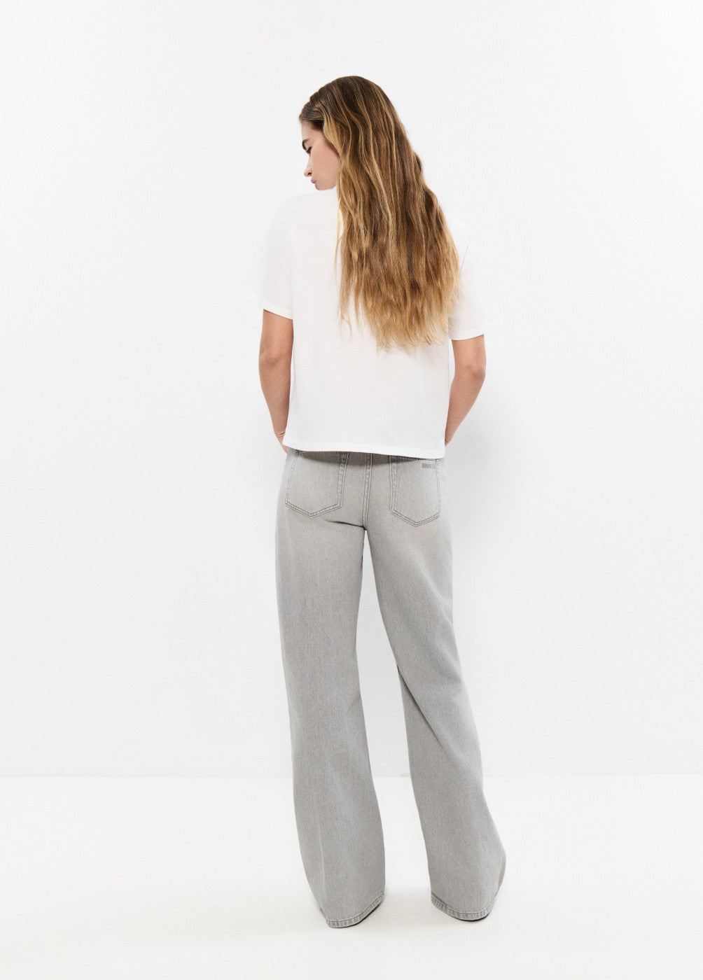 אישה עם חולצה לבנה ומכנסי ג'ינס אפורים רחבים, עומדת עם גב למצלמה על רקע לבן