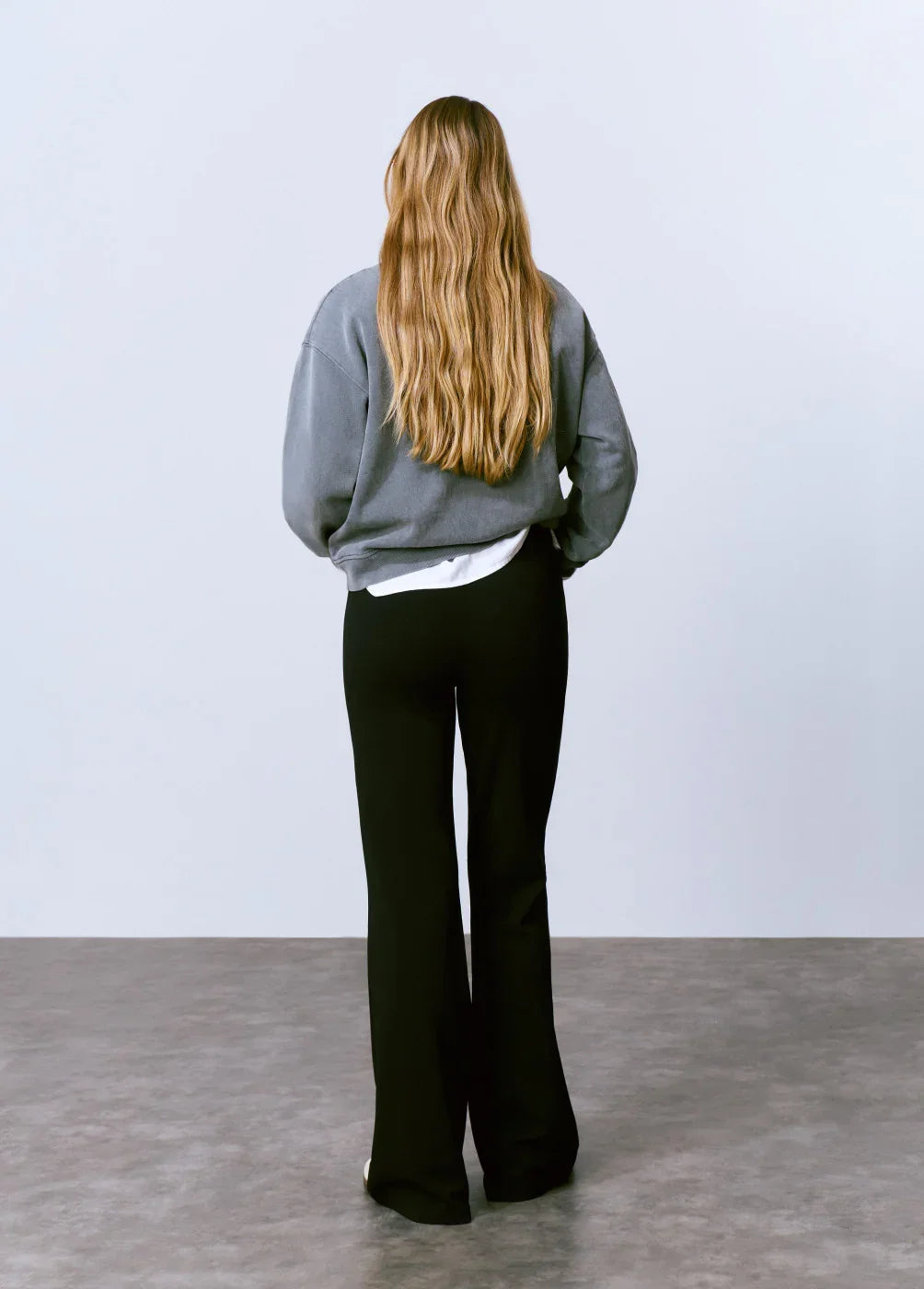 אישה עם שיער בלונדיני גלי, לבושה בסווטשירט אפור ומכנסיים שחורים, עומדת על רצפת בטון רקע לבן, מותגים מובילים CHOZEN