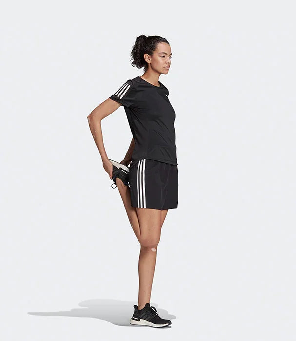 אישה בספורט, לובשת חולצת ושורטס אדידס שחורים עם פסים לבנים, מדגימה מתיחה, רקע אפור