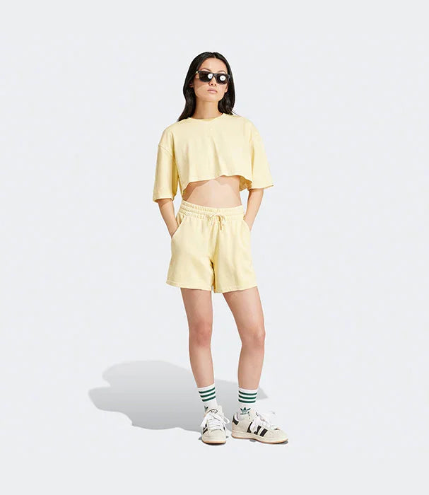 אישה לובשת סט קיץ צהוב, טי שירט קצרה ומכנסיים קצרים, משקפי שמש ונעלי אדידס, צילום על רקע אפור ל-CHOZEN