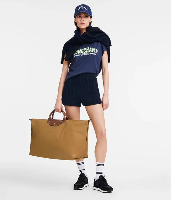 אישה בלבוש ספורטיבי עם כובע כחול, חולצת LONGCHAMP, מכנסיים קצרים ותיק גדול חום. מוצר אופנה יוקרתי ב-CHOZEN.