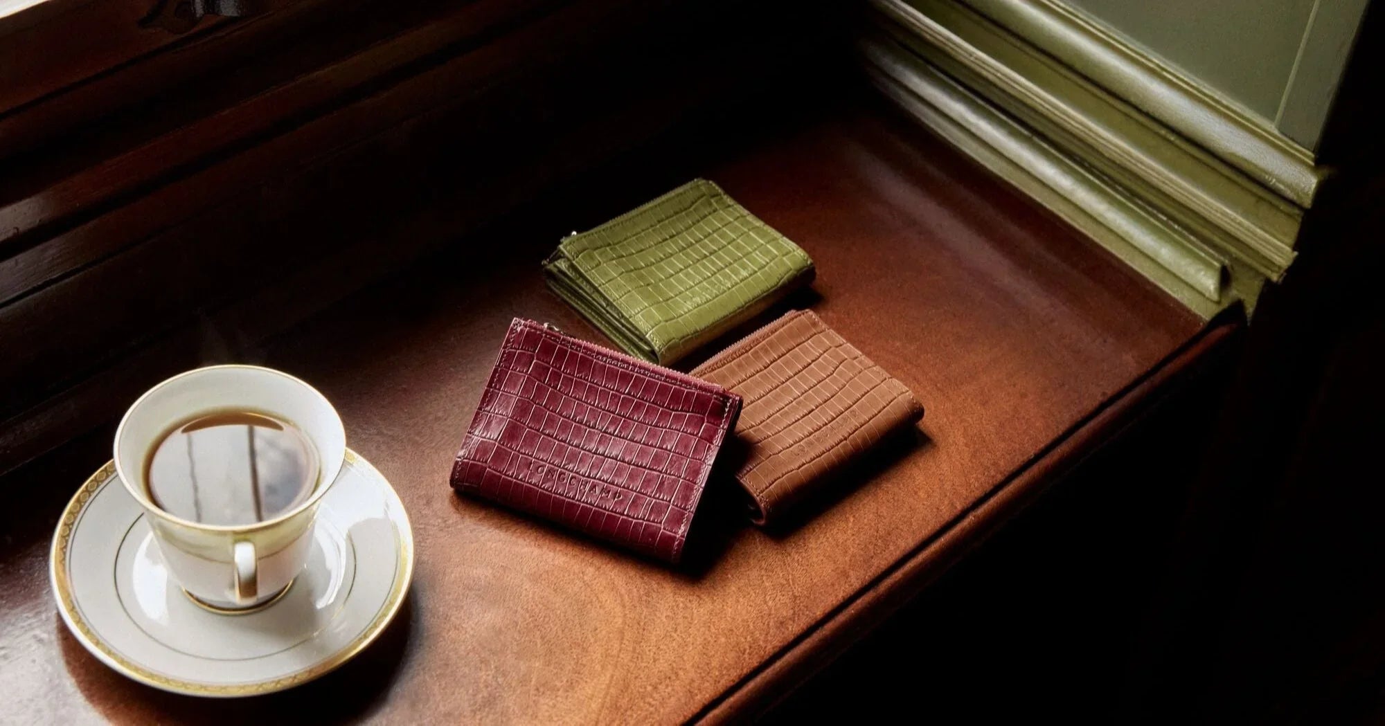 ארנקים יוקרתיים בצבע ירוק, חום ובורדו ליד כוס קפה על שולחן עץ, CHOZEN מותגים מובילים