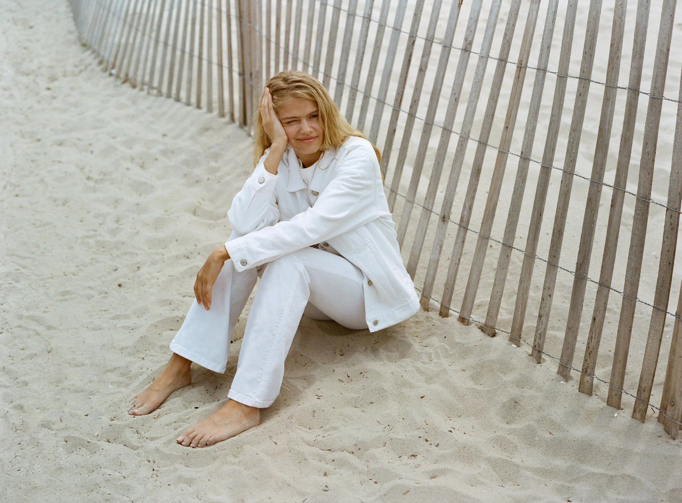 אישה בלבוש לבן יושבת על חול ליד גדר עץ בחוף, אופנה יוקרתית CHOZEN