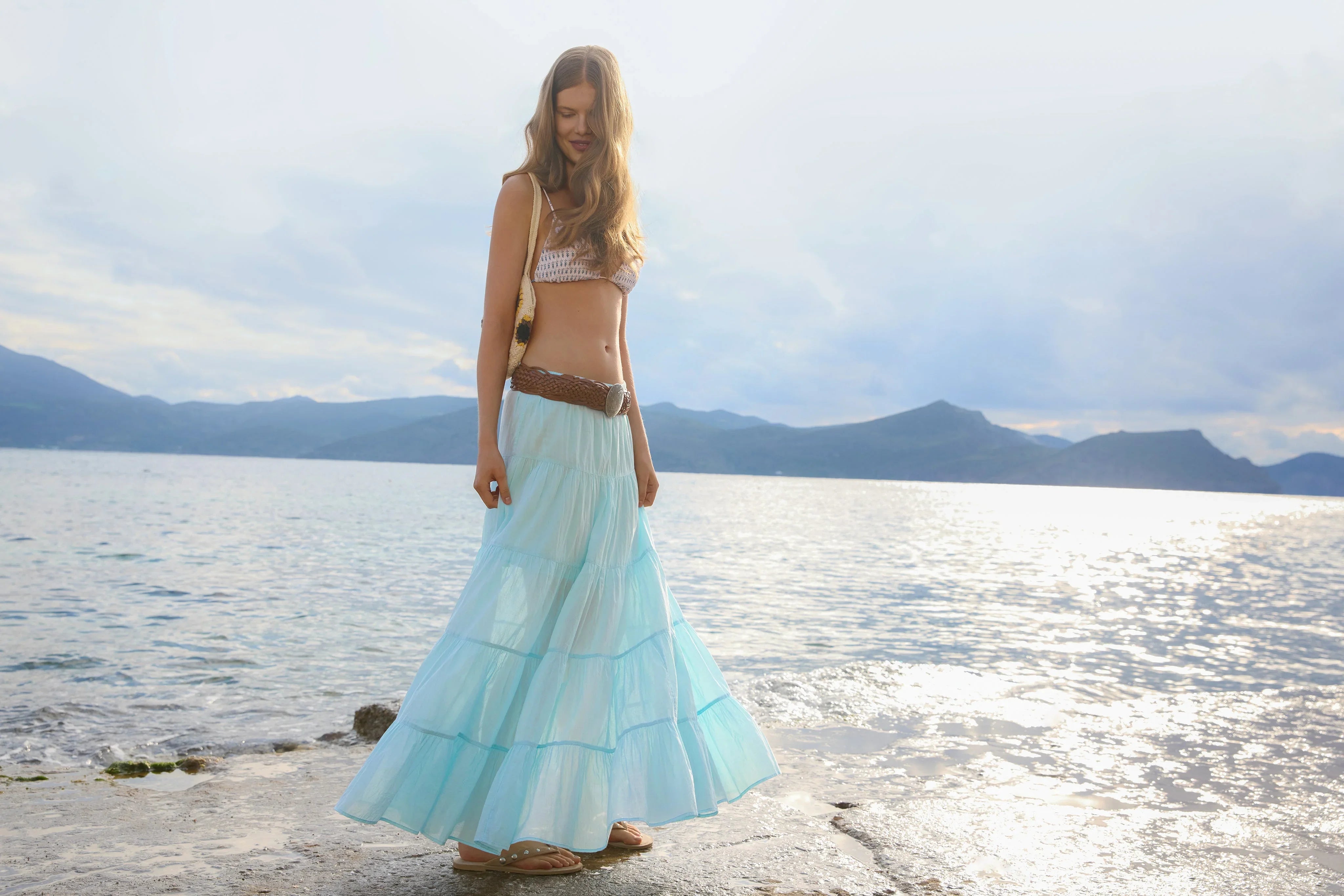 דוגמנית בשמלה ארוכה תכולה על שפת ים, אופנה קיץ CHOZEN, רקע הרים ומים