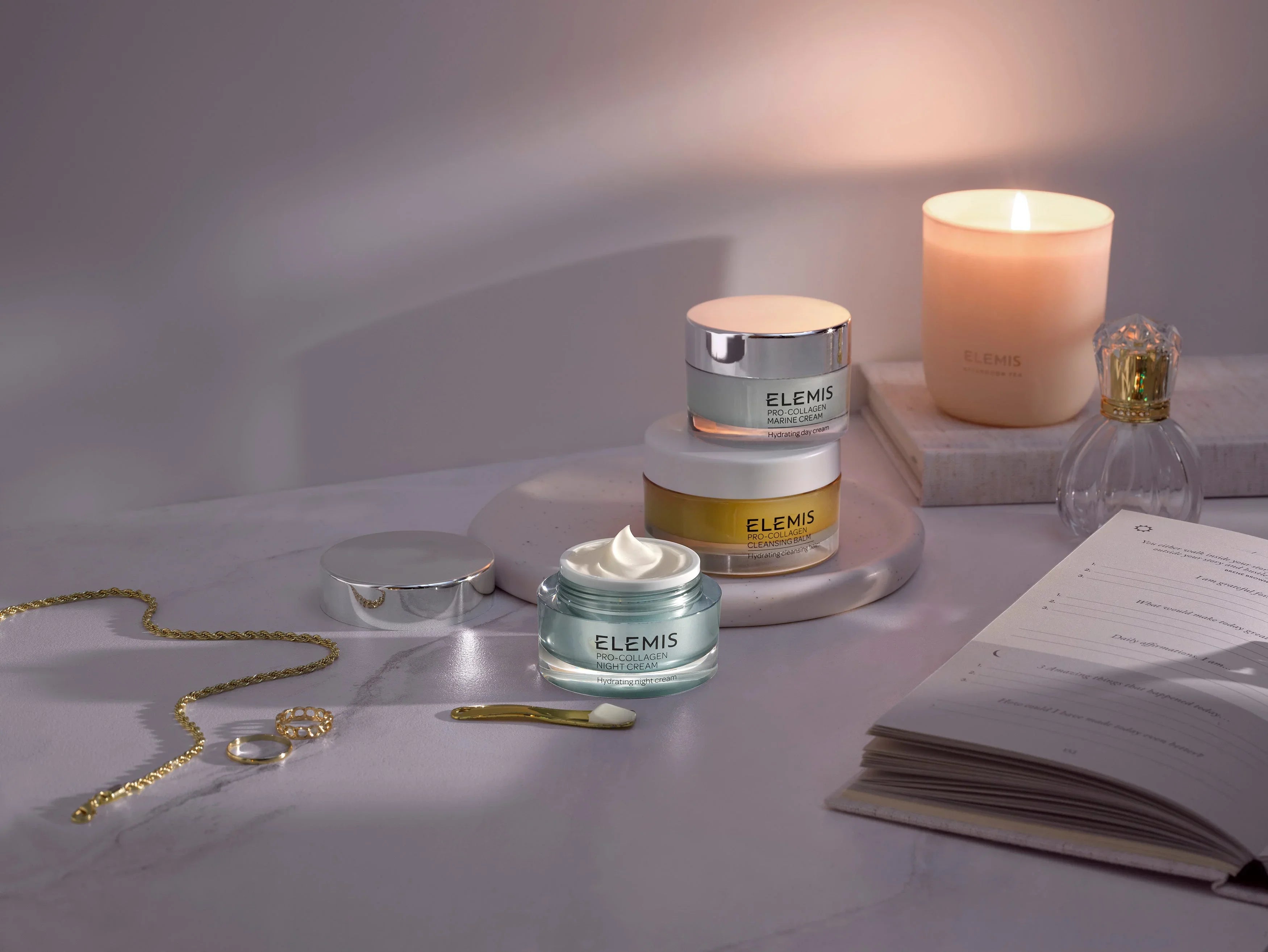 קרם פנים ELEMIS על שולחן שיש לצד נר דולק, תכשיטים וספר, מוצג באתר CHOZEN
