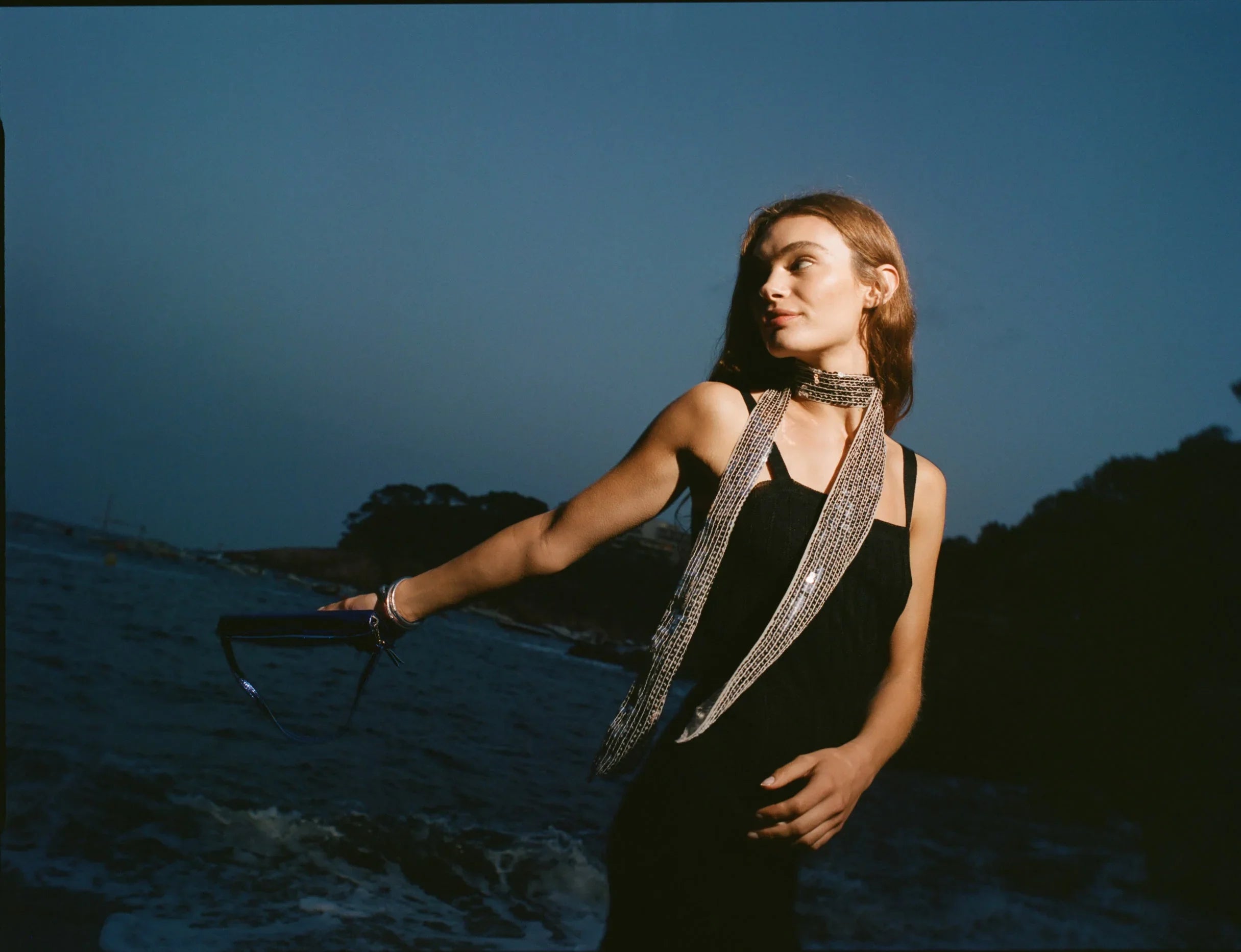 אישה בשמלה שחורה וצעיף נוצץ על רקע חוף ים בשקיעה, אופנה יוקרתית CHOZEN