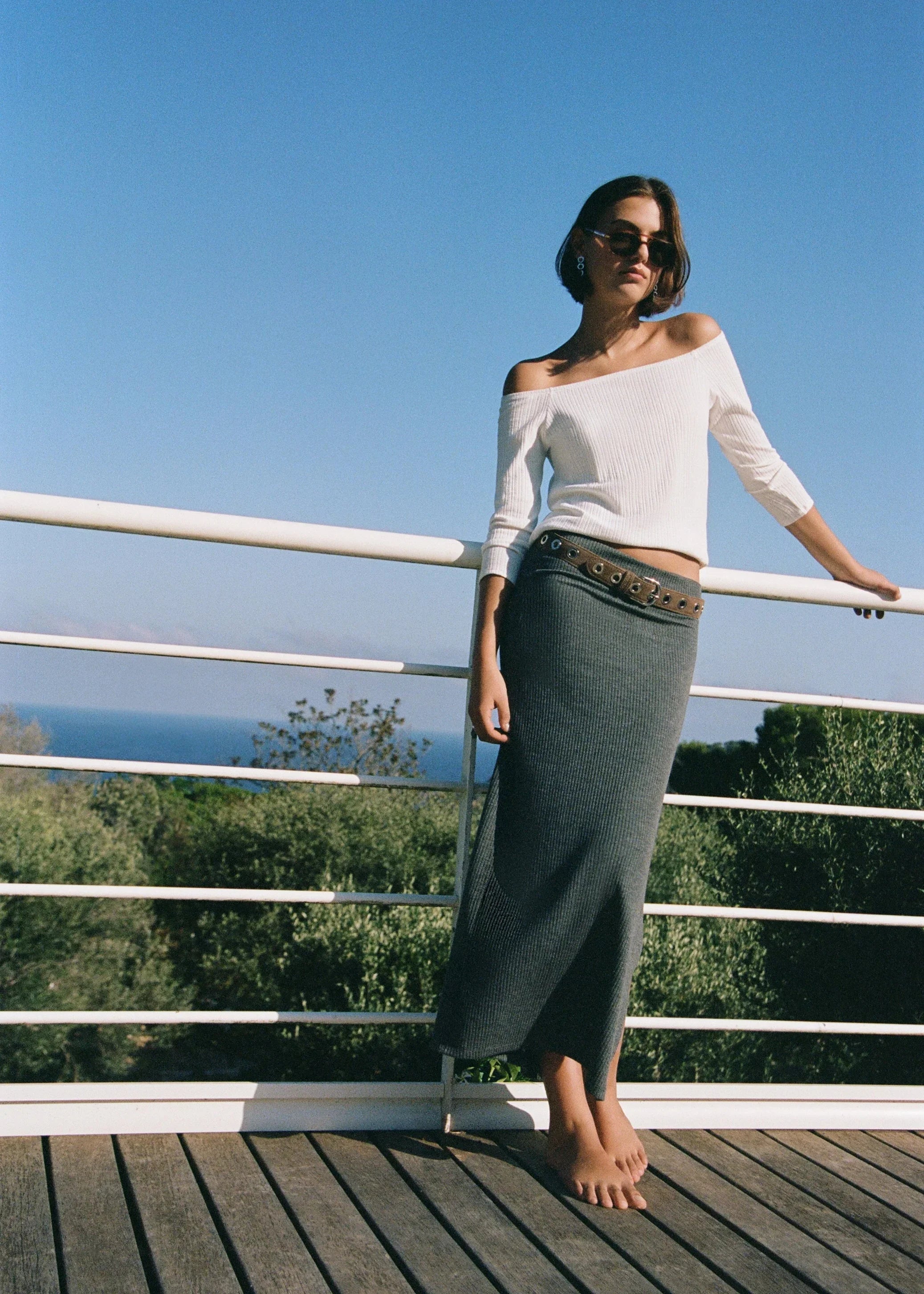 אישה בטרסה עם נוף לים, לובשת חולצה לבנה עם כתף חשופה וחצאית ירוקה ארוכה, אופנה CHOZEN