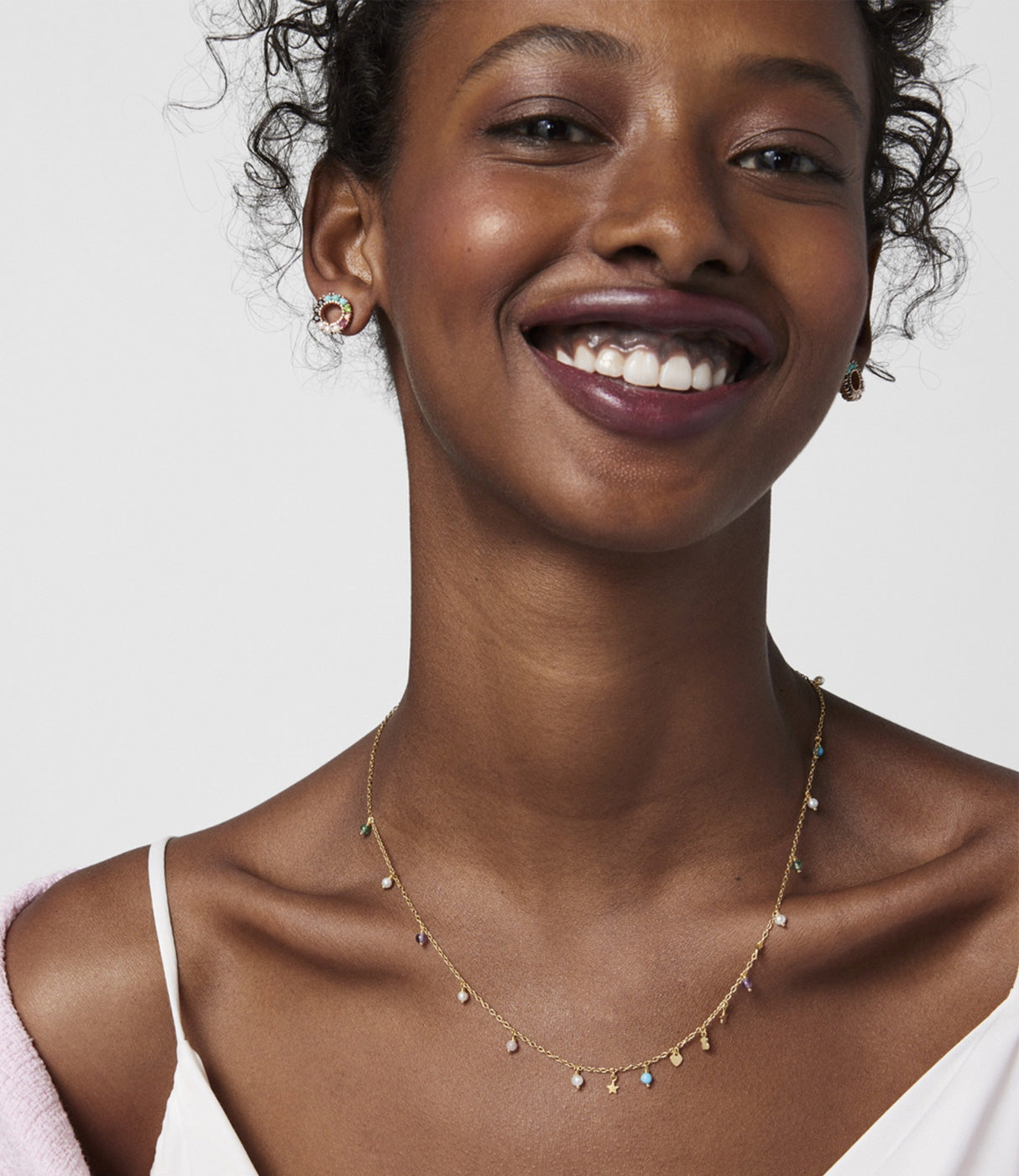 אישה מחייכת עונדת שרשרת זהב עדינה עם תליונים צבעוניים ועגילי זהב, תכשיטי אופנה אלגנטיים