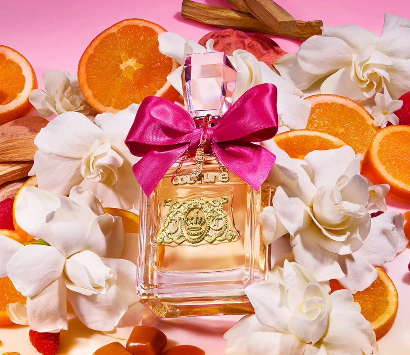 בקבוק בושם עם פקק קריסטל וסרט ורוד, מוקף בפרחי גרדן, פרוסות תפוז וקוביות קרמל