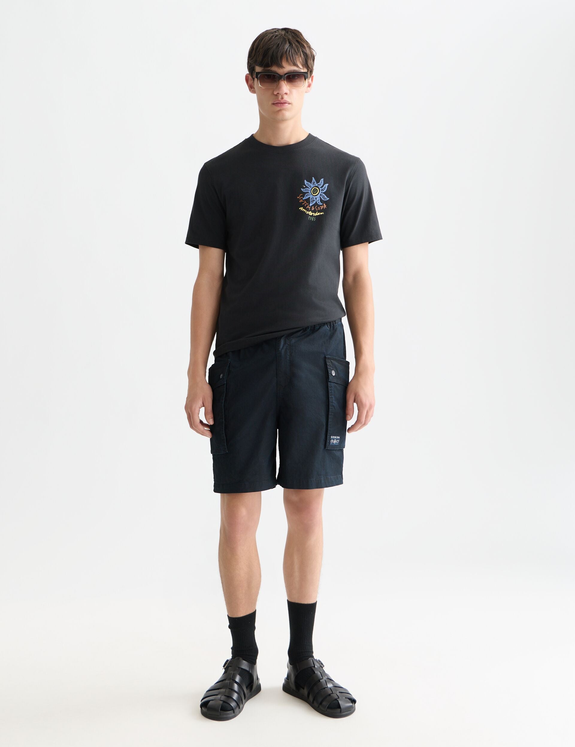 דגם גבר לובש חולצת טי שחורה עם לוגו, מכנסיים קצרים שחורים ונעלי סנדל טרנדיות, CHOZEN בגדי מותגים