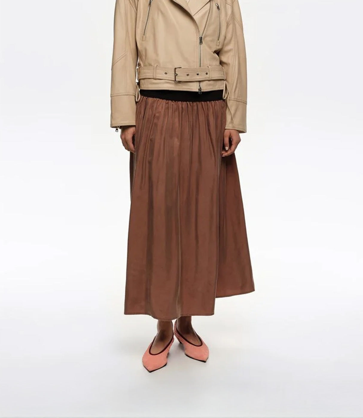 אישה לבושה בז'קט עור בז', חצאית ארוכה חומה ונעליים ורודות, רקע לבן