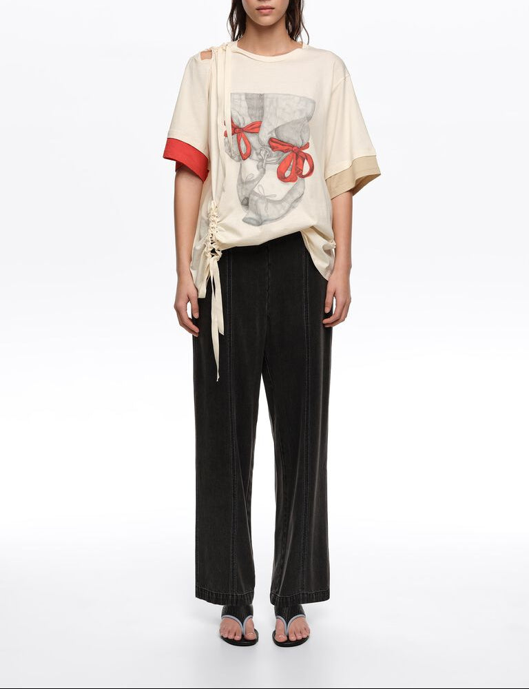 אישה לובשת חולצת טי בהירה עם הדפס גרפי וקשרי סרט אדומים, מכנסיים שחורים רחבים וסנדלים