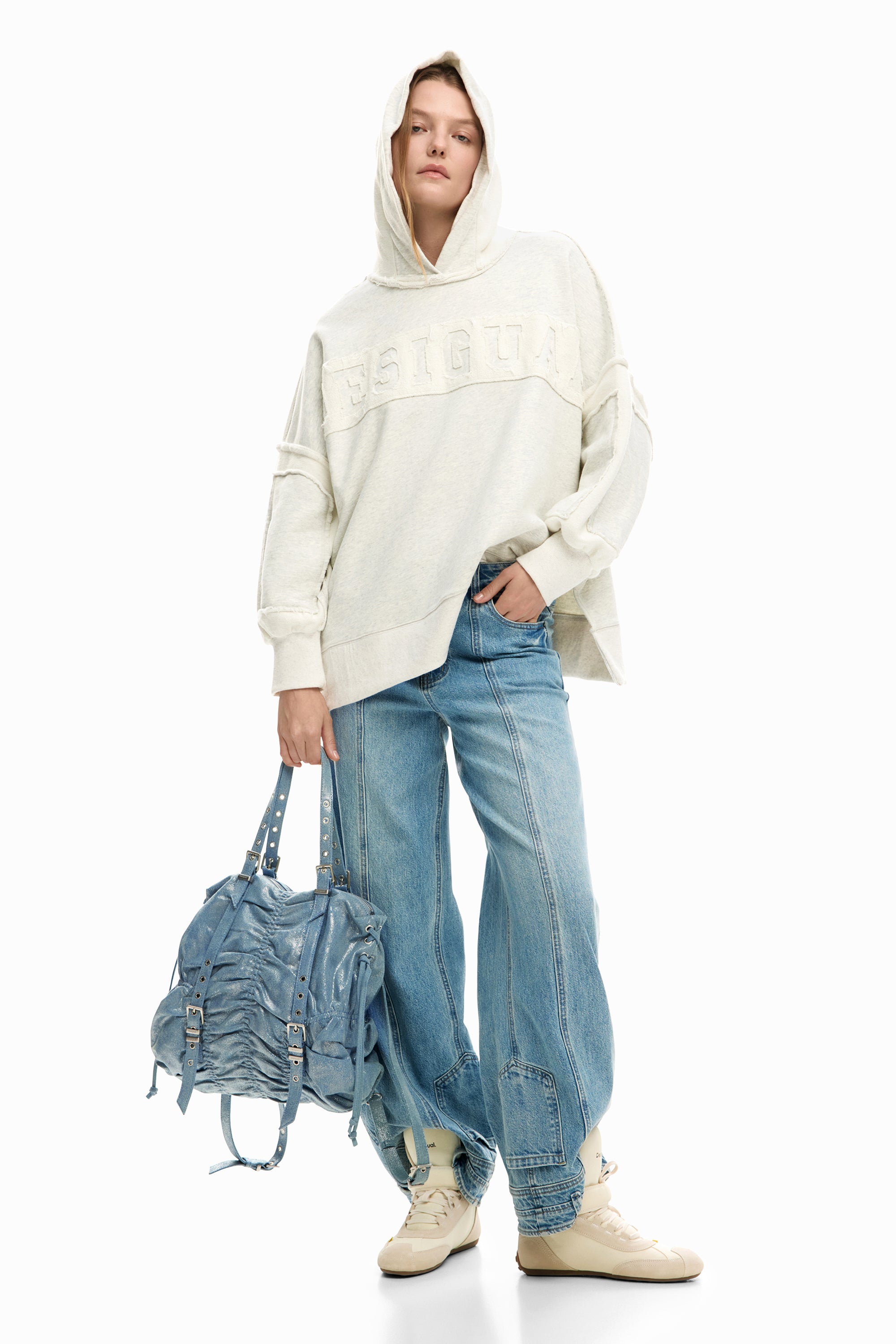 דוגמנית עם סווטשירט קפוצ'ון קרם, ג'ינס כחול רחב, נעליים בהירות ותיק יד גדול בגוון ג'ינס