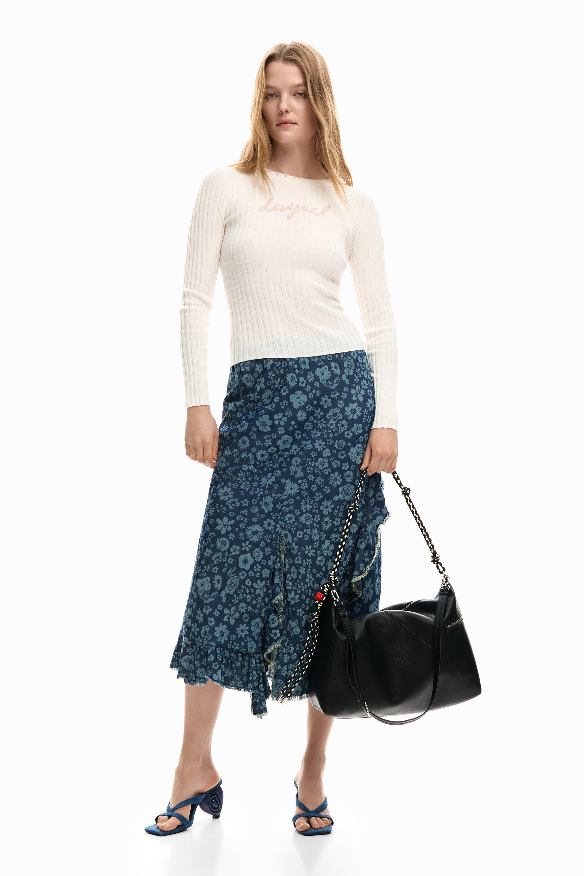 אישה עם חצאית פרחונית כחולה, סוודר לבן, תיק כתף שחור ונעלי עקב כחולות