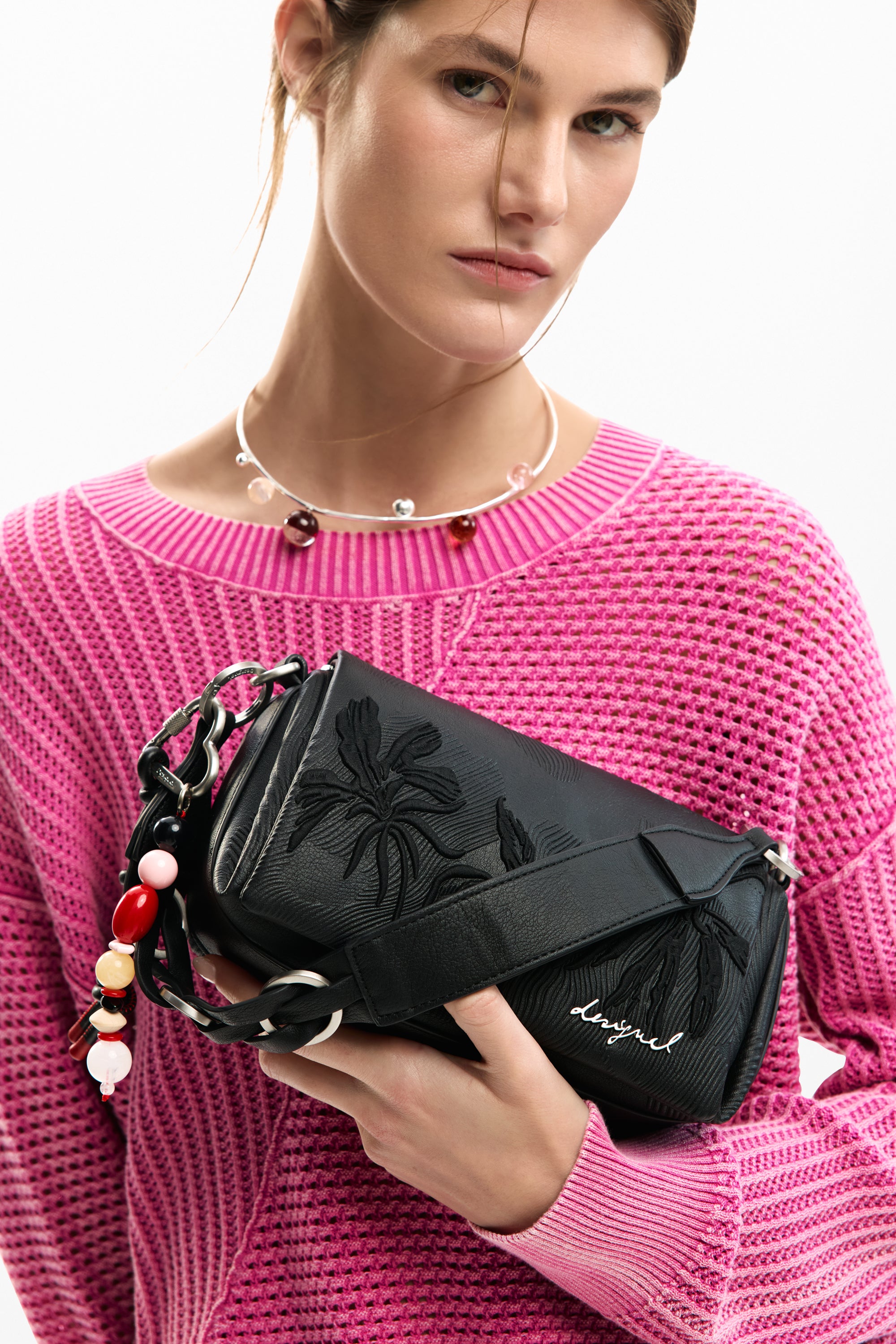 אישה בסוודר ורוד מחזיקה תיק צד שחור עם עיטורי פרחים ושרשרת חרוזים צבעונית