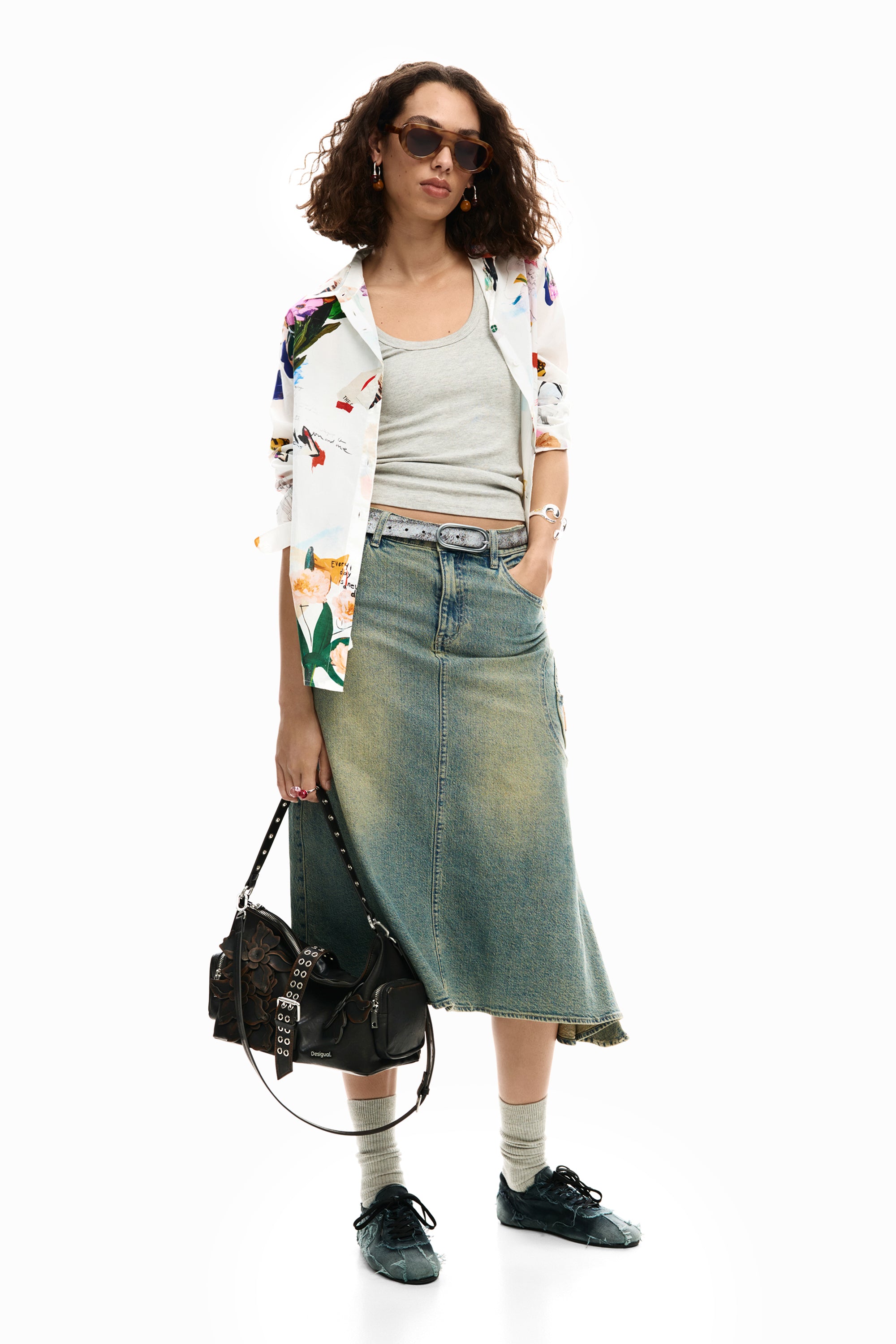אישה עם חולצה פרחונית, גופיה אפורה, חצאית ג'ינס ארוכה, נעלי סניקרס, תיק שחור ומשקפי שמש אופנתיים