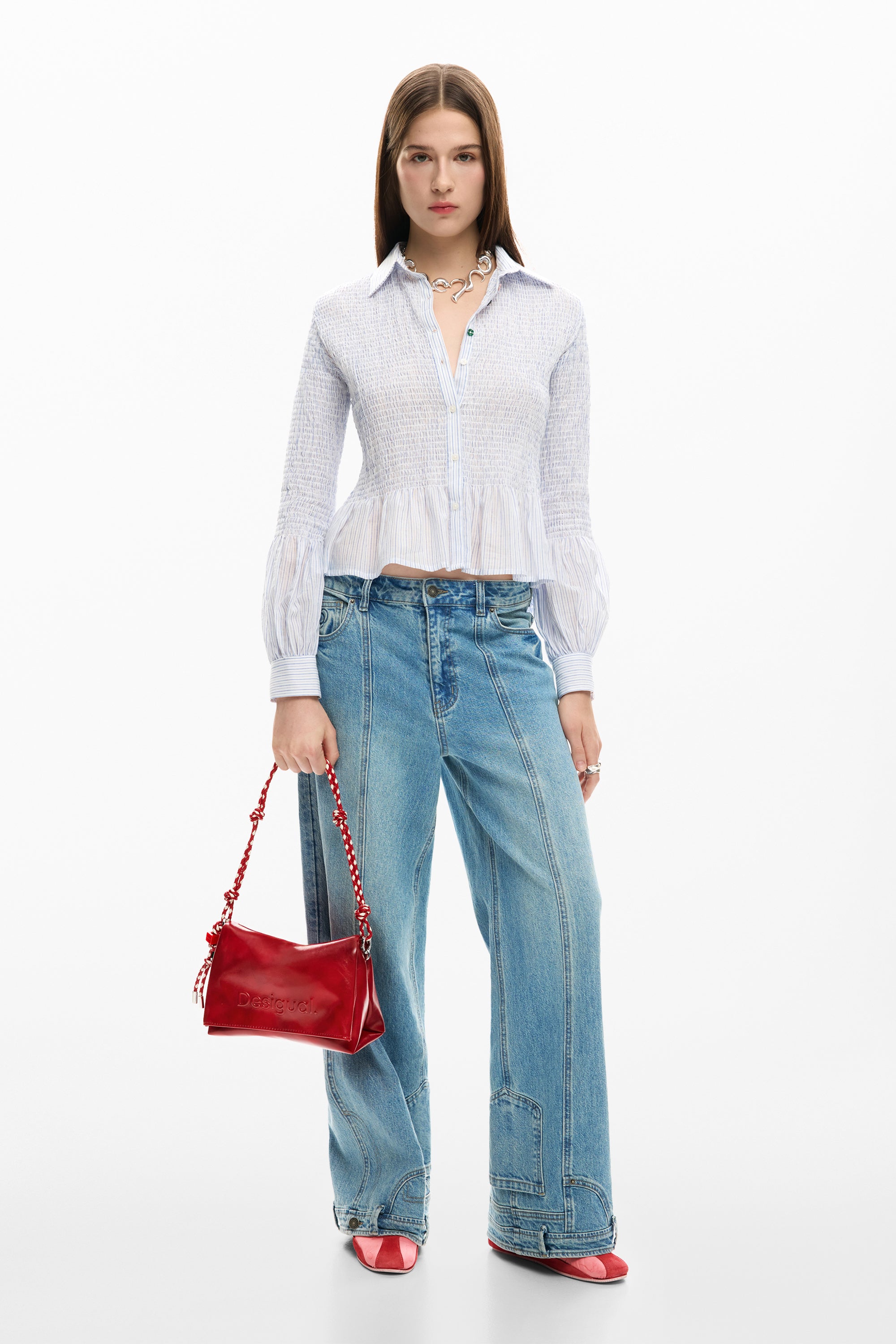 אישה עם חולצה לבנה מכופתרת, מכנסי ג'ינס רחבים, תיק אדום ונעלי עקב אדומות, רקע לבן