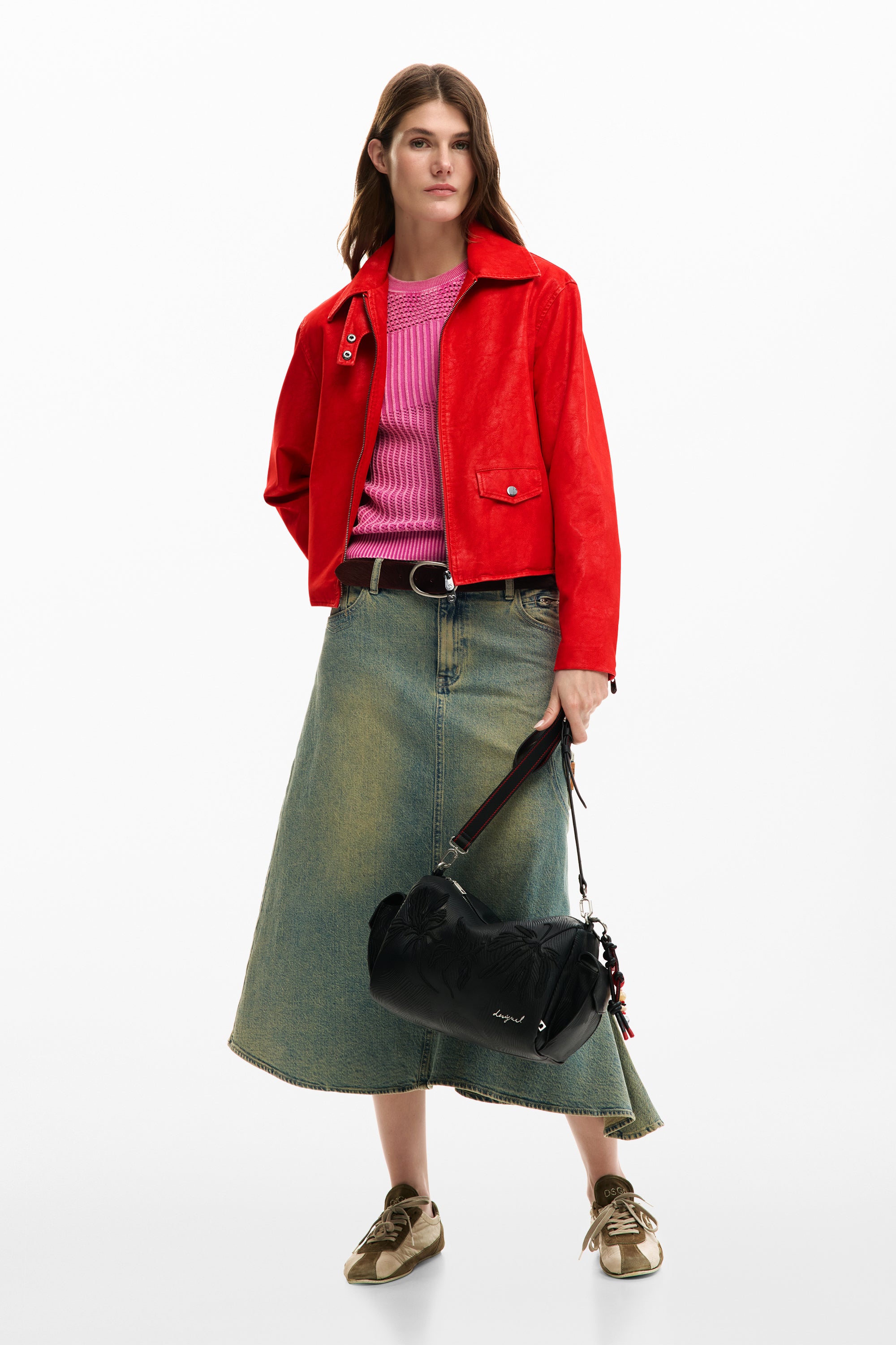 אישה עם ז'קט אדום, חולצה ורודה, חצאית ג'ינס ותיק צד שחור אופנתי, עומדת בסטודיו