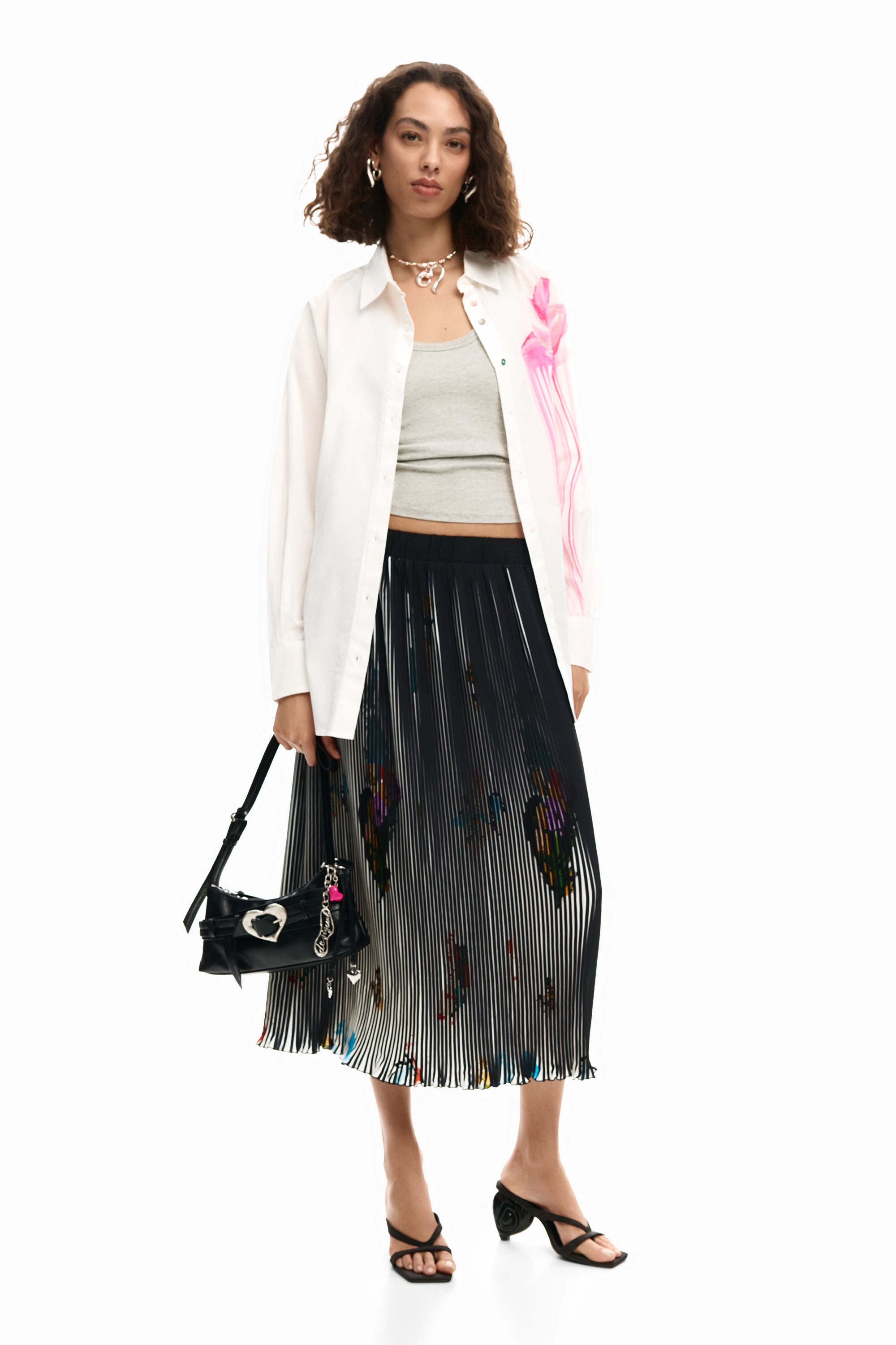 אישה עם חולצה לבנה עם הדפס ורוד, גופיה אפורה, חצאית פליסה שחורה תיק קטן ונעלי עקב