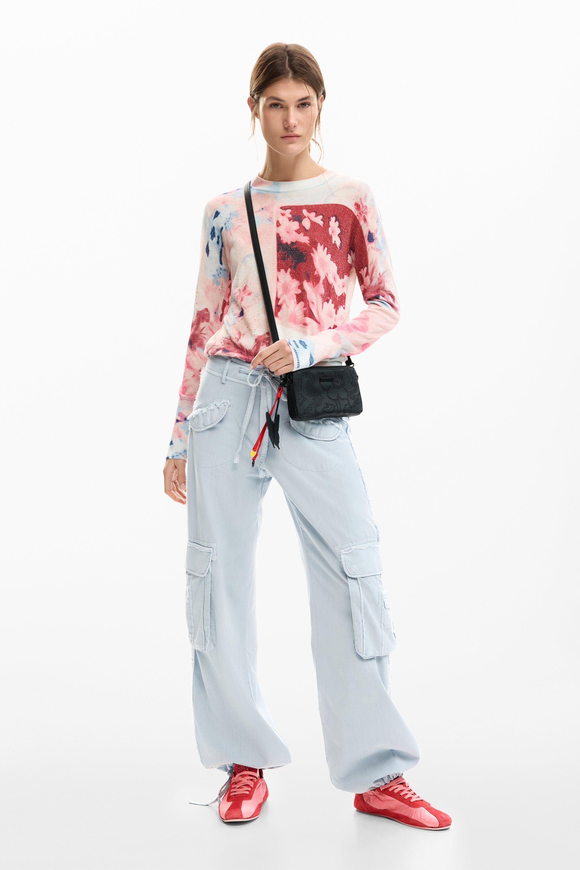 אישה עם סווטשירט פרחוני, מכנסי קארגו בהירים, נעלי ספורט אדומות ותיק כתף שחור