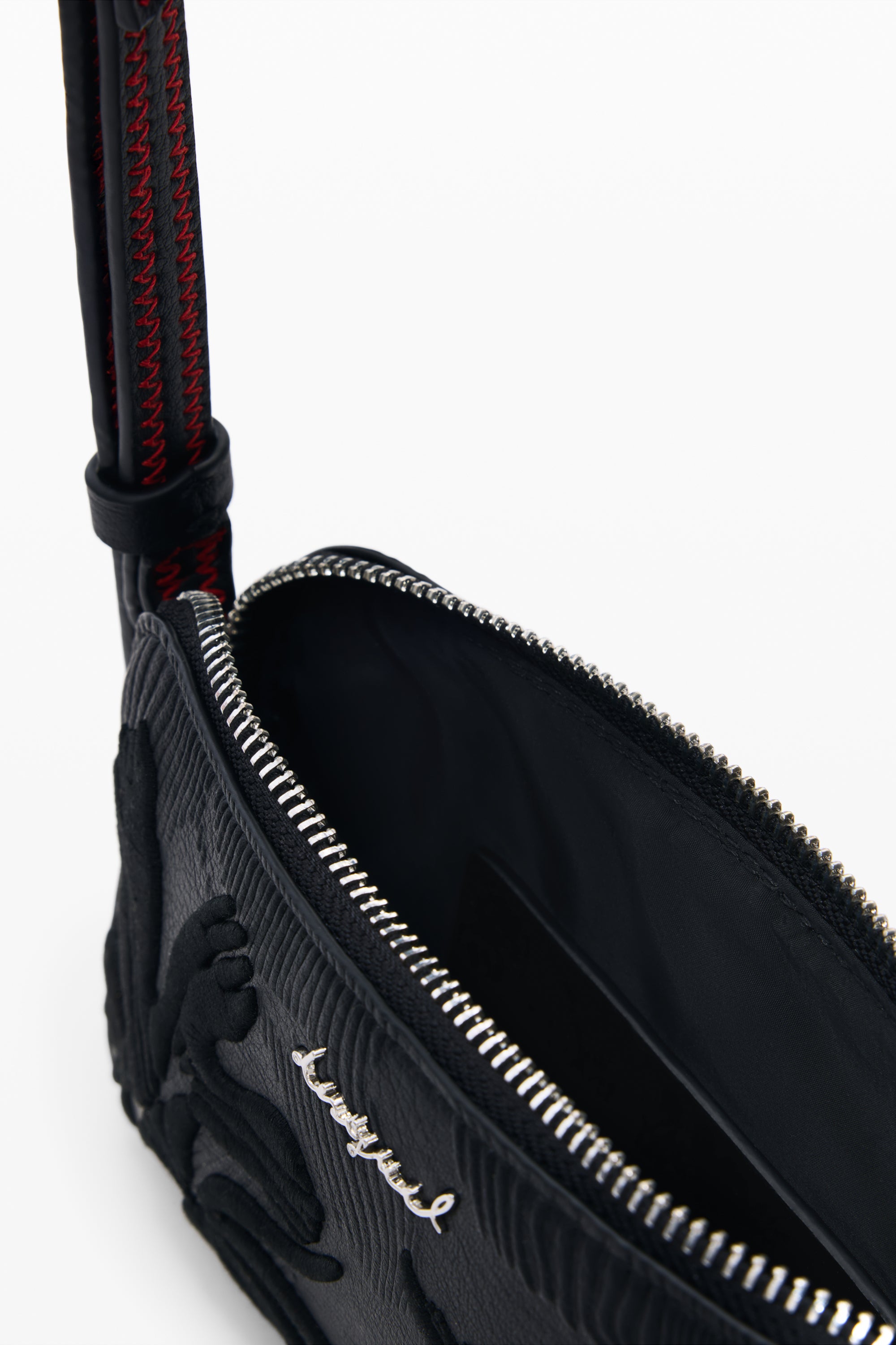 תצלום קלוז-אפ של תיק צד קטן שחור עם רוכסן פתוח, רקמת עיטור וכיתוב מתכתי, רצועה עם תפרים אדומים.