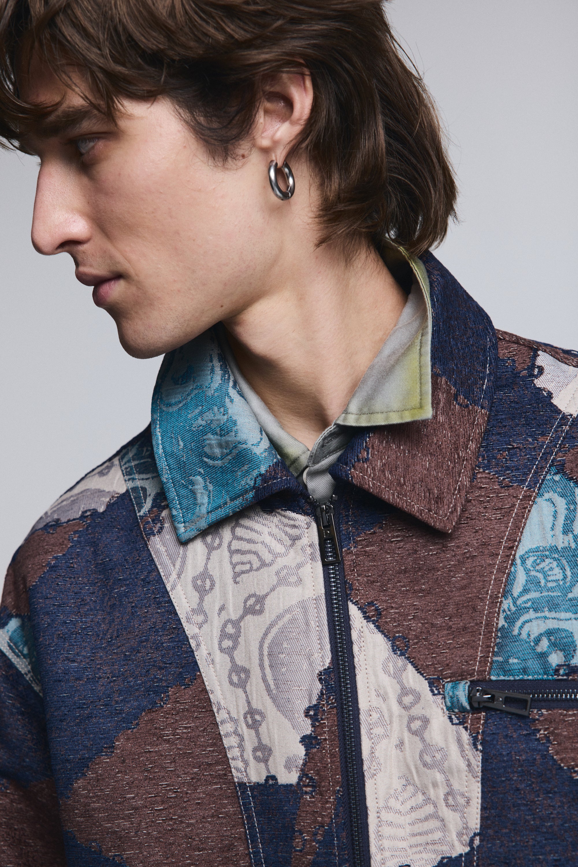 גבר צעיר עם עגיל כסף לובש ז'קט ג'קארד צבעוני, דוגמת פסיפס בצבעי כחול, חום ובז'