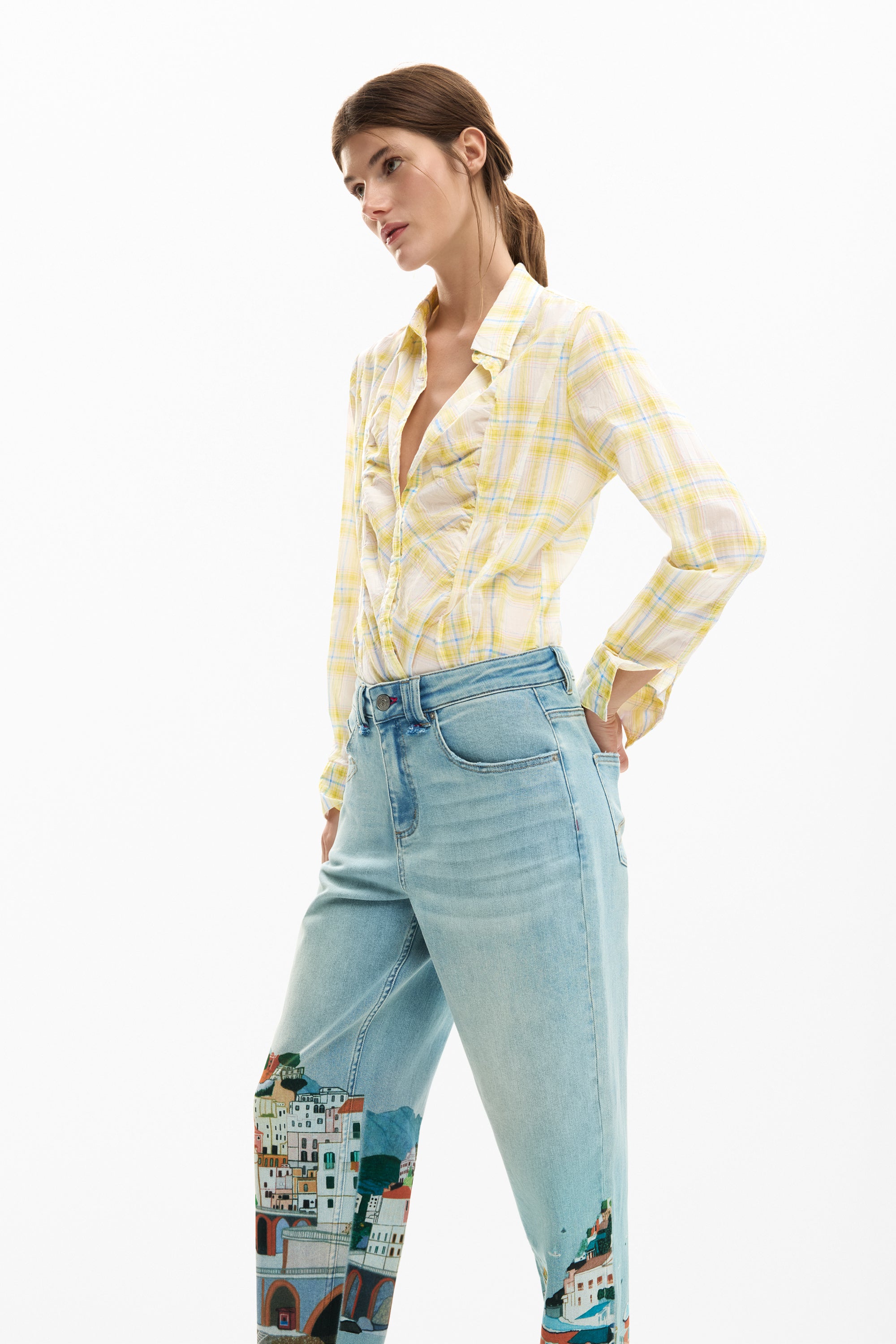 אישה לובשת חולצת משבצות צהובה ומכנסי ג׳ינס בהירים עם הדפס בניינים צבעוני על הרגליים