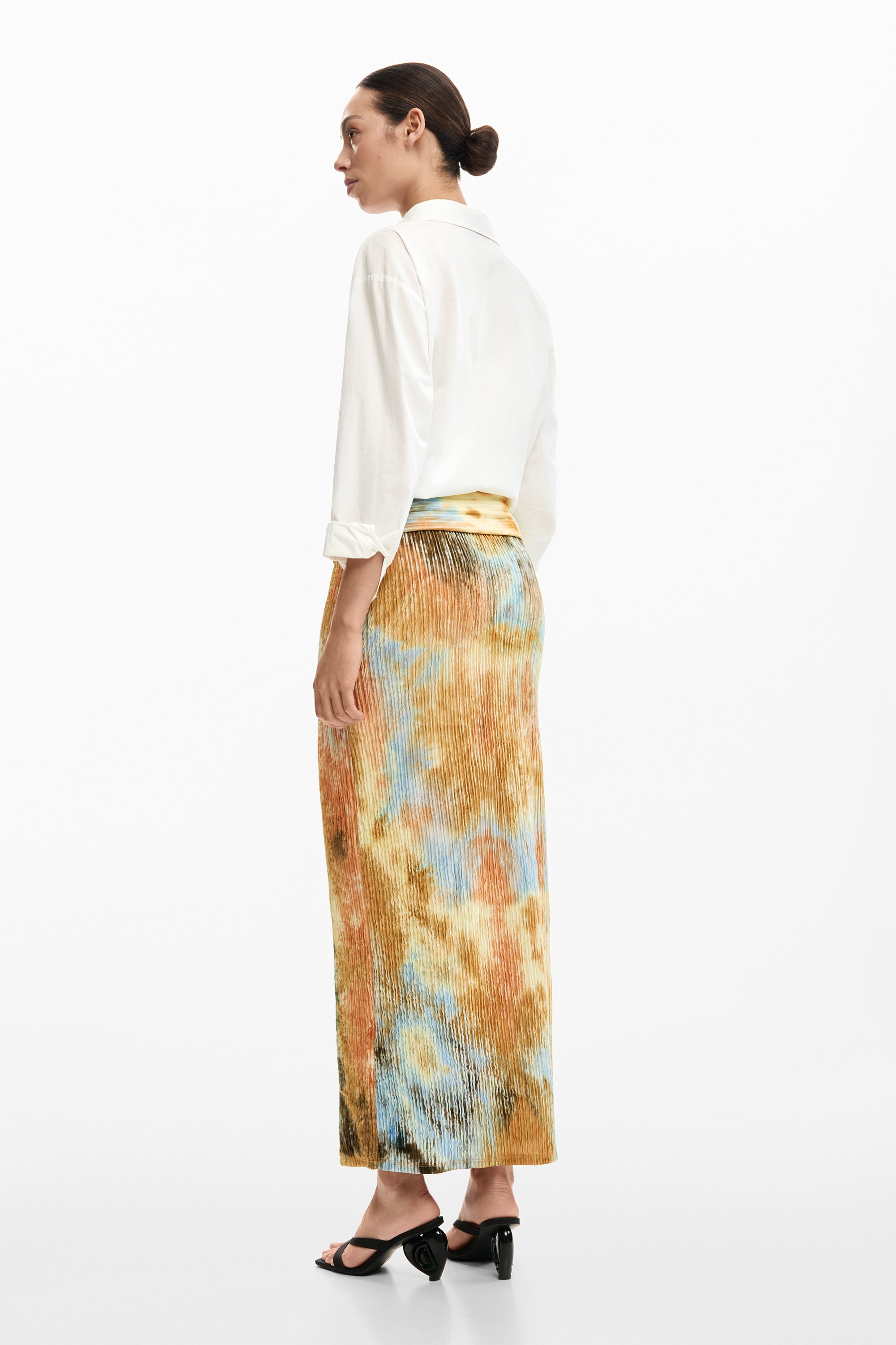 אישה לובשת חצאית פליסה צהובה-כתומה בהדפס ססגוני עם חולצה לבנה וכפכפי עקב שחורים