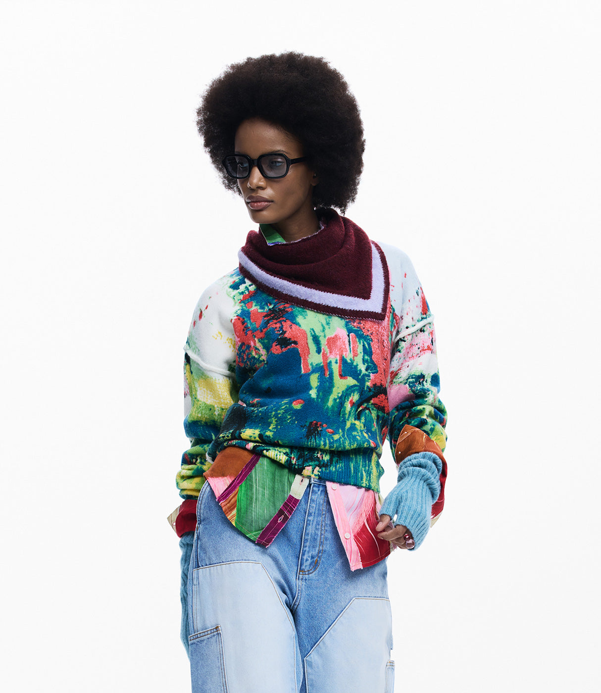 אישה עם שיער אפרו, לובשת סוודר צבעוני ודנים, צעיף בורדו ומשקפי אופנה עגולים