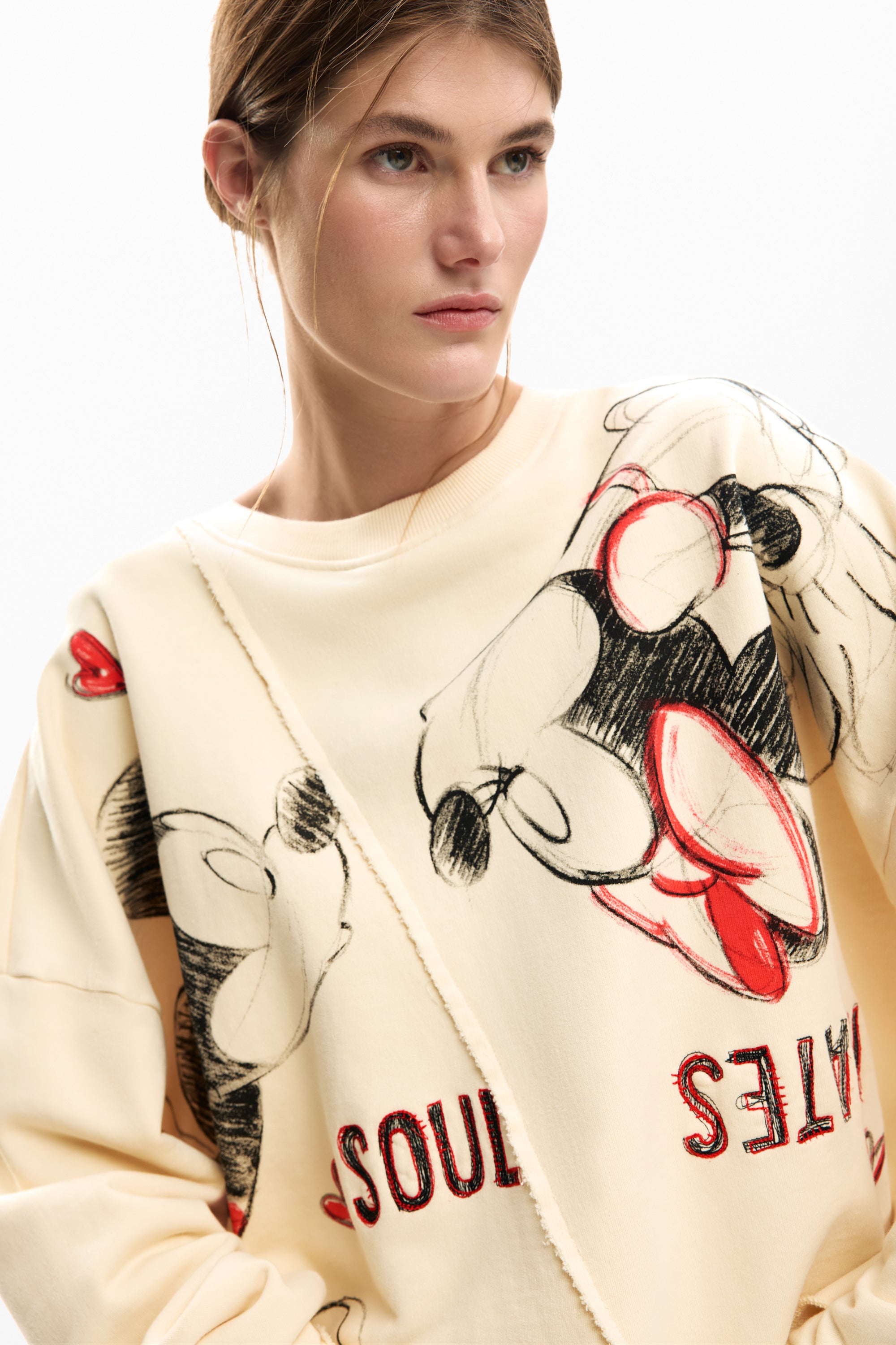 אישה לובשת סווטשירט קרם עם הדפס דמויות קומיקס צבעוניות ומילה SOULMATES באדום