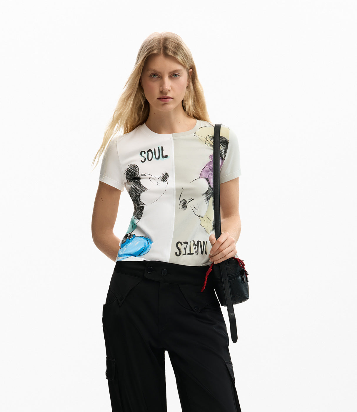 אישה לובשת חולצת טי מודפסת עם דמויות מצוירות וצבעים שונים, מכנסיים שחורים ותיק צד קטן