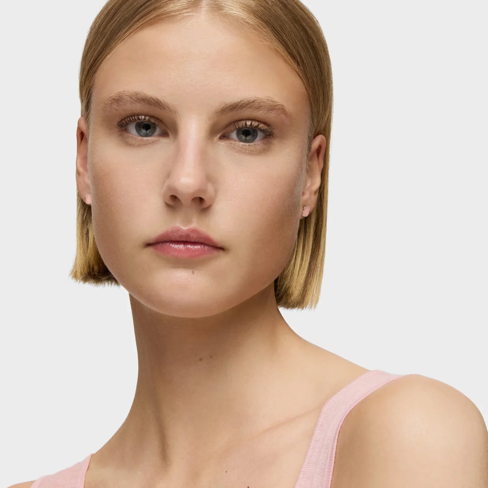 אישה בלונדינית עם שיער קצר עונדת עגילים קטנים, לובשת גופיה ורודה, רקע לבן