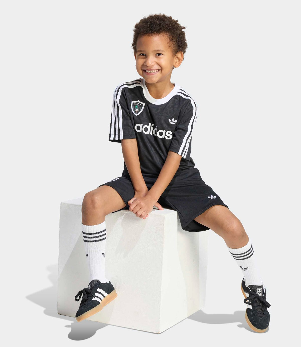ילד יושב על קובייה לבנה לובש סט בגדי ספורט שחור עם פסים לבנים ונעליים תואמות