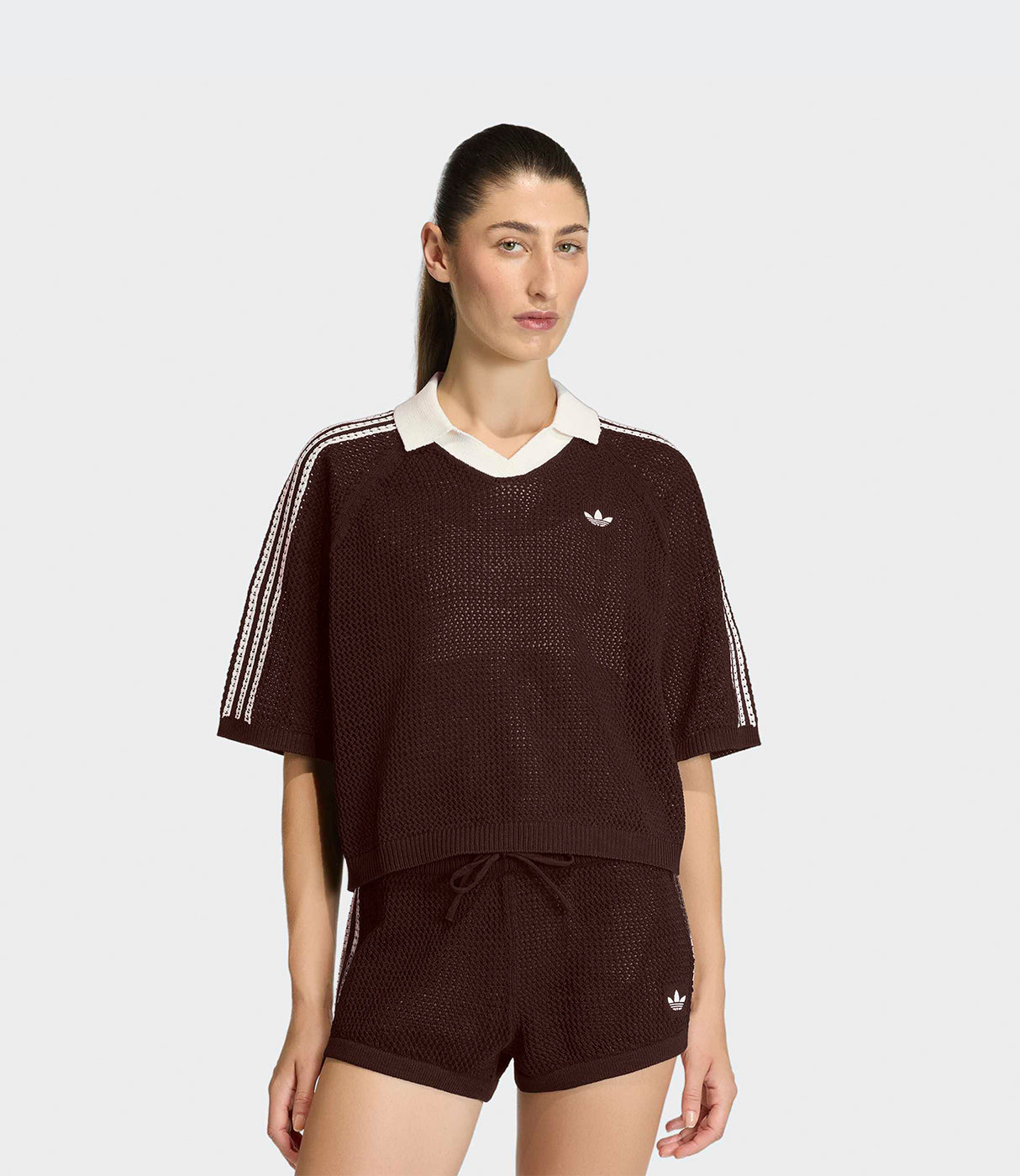 אישה לובשת סט בגדי ספורט חומים קצרים עם פסים לבנים וסמל אדידס, רקע בהיר