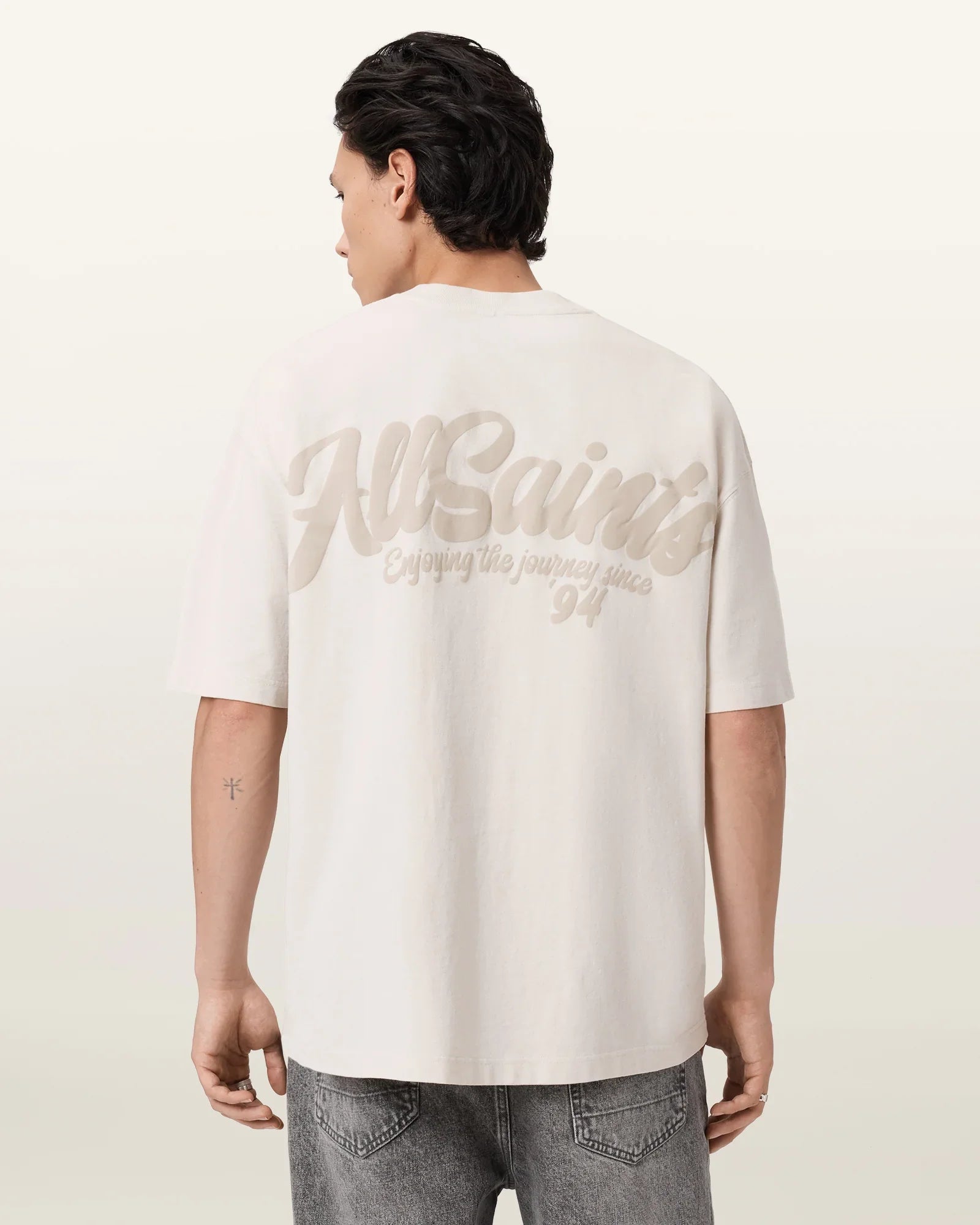 גבר לובש חולצת טי שירט לבנה עם הדפס AllSaints בגב, ג'ינס אפור, רקע בהיר