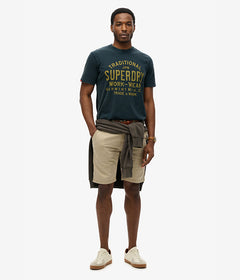גבר לובש חולצת טי Superdry כחולה, מכנסי ברמודה בז', נעלי סניקרס לבנות וסווטשירט קשור במותן