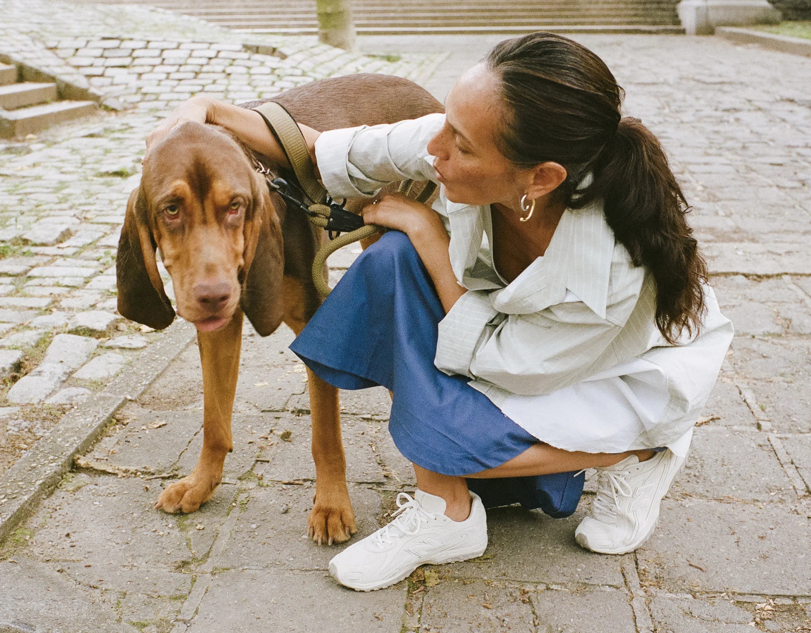 אישה בחצאית כחולה חולצת פסים ונעלי סניקרס לבנות עם כלב גדול ברחוב מרוצף, CHOZEN
