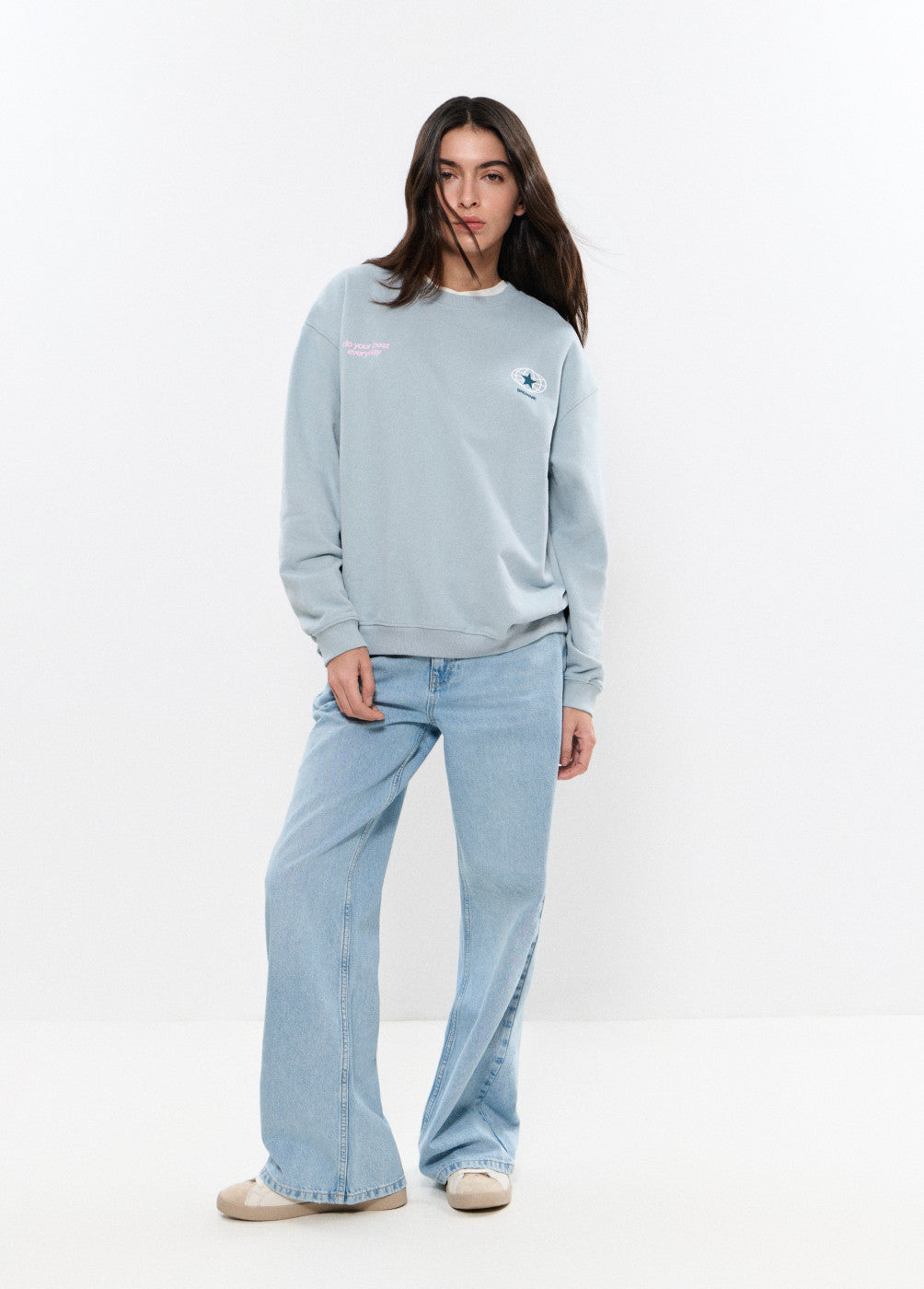 אישה לובשת סווטשירט תכלת עם כיתוב ולוגו, ג'ינס בהיר רחב ונעלי בד בז', סטייל קז'ואל