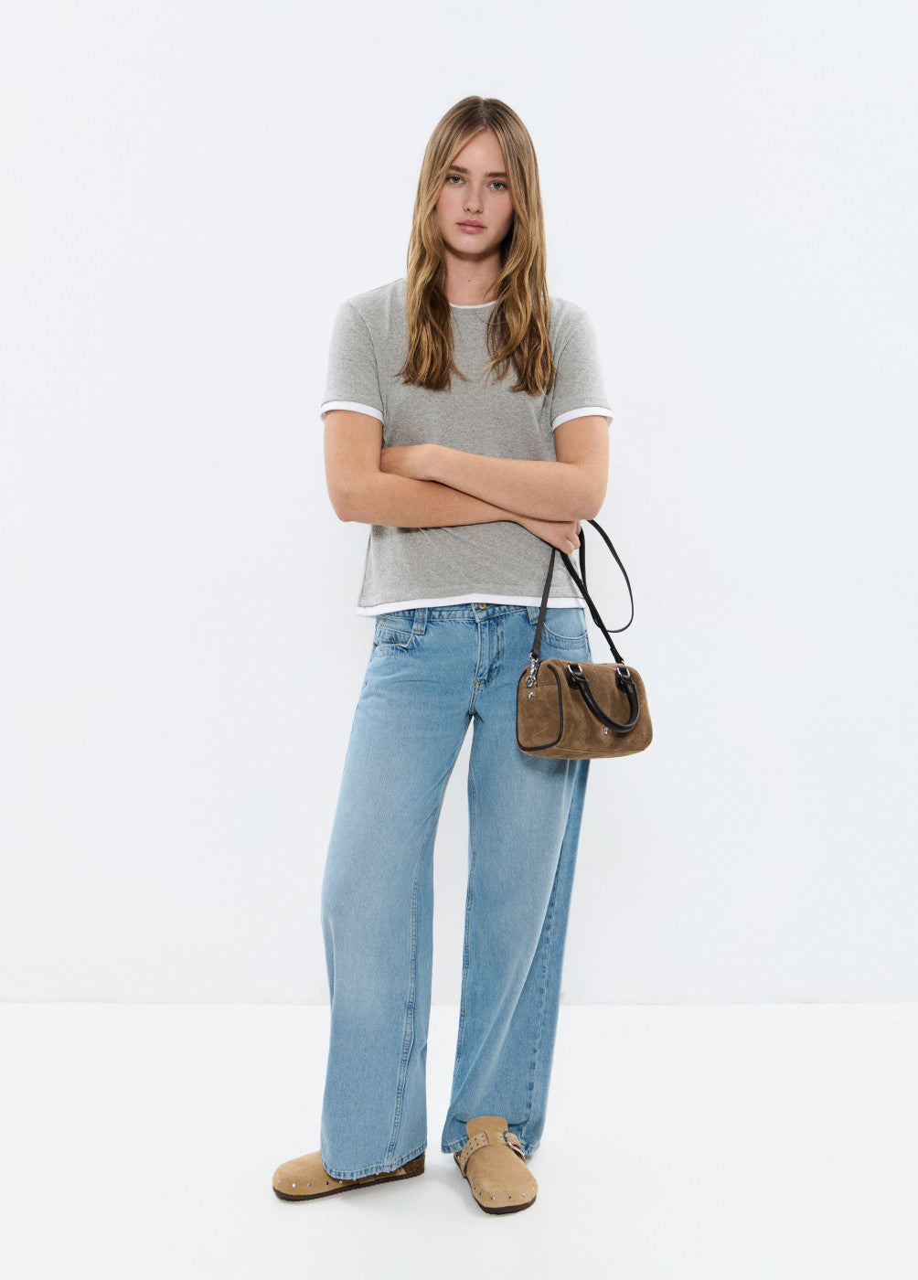 אישה צעירה לובשת חולצת טי אפורה, ג'ינס רחב ותיק חום בעמידה על רקע לבן