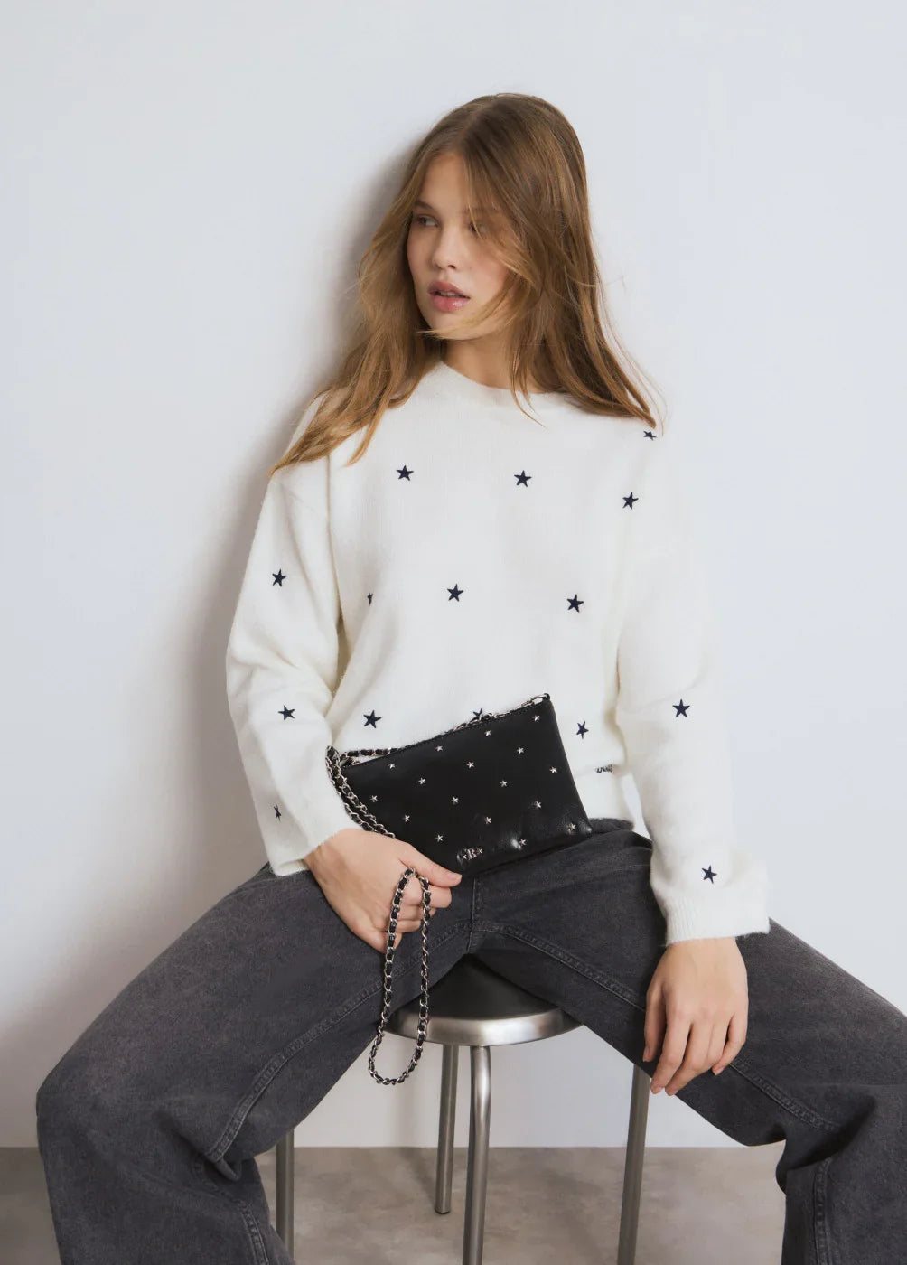 דוגמנית לובשת סוודר לבן עם כוכבים שחורים ומחזיקה תיק קלאץ' שחור עם שרשרת, אופנה CHOZEN