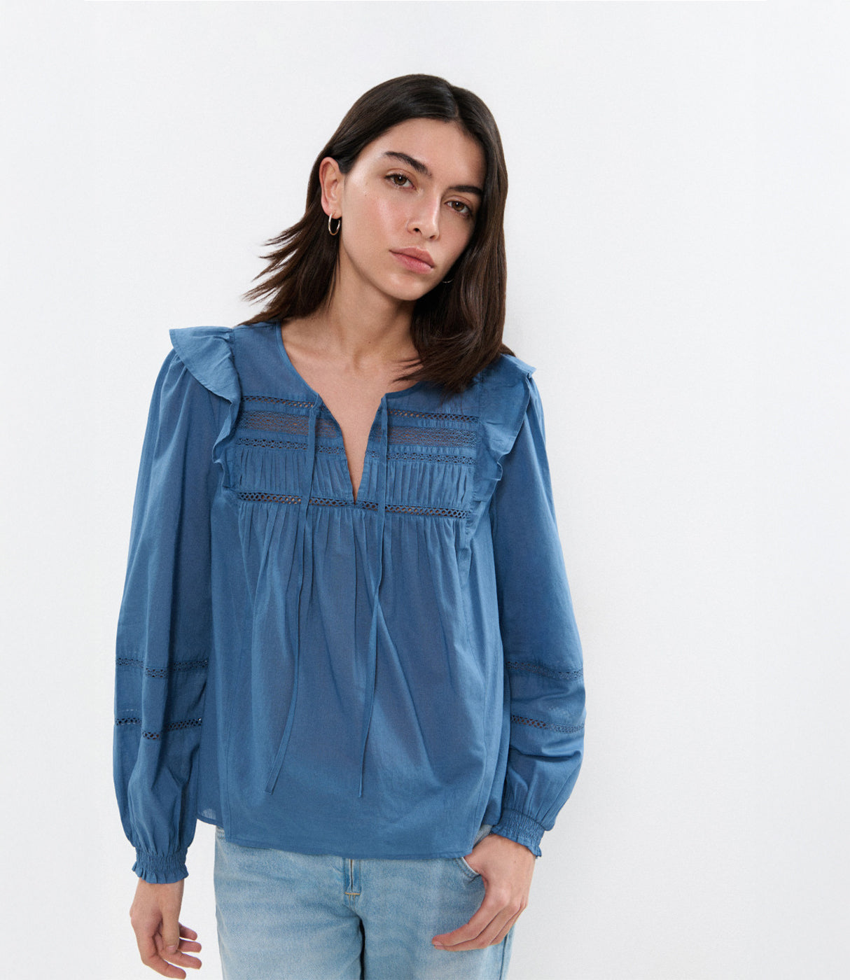 אישה לובשת חולצה כחולה ארוכה עם שרוולים תפוחים ותחרה, מכנסי ג'ינס בהירים, רקע לבן