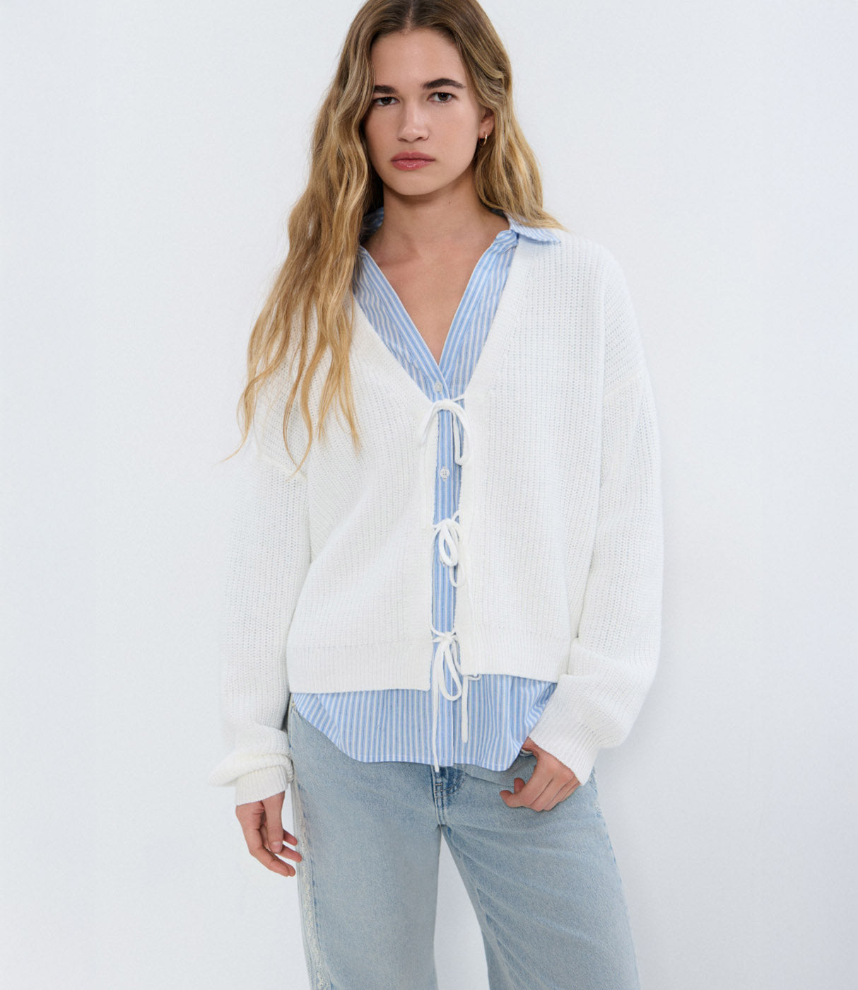 אישה לובשת סוודר לבן קליל עם שרוכים מעל חולצה מכופתרת כחולה מפוספסת ומכנסי ג'ינס, אופנת נשים טרנדית